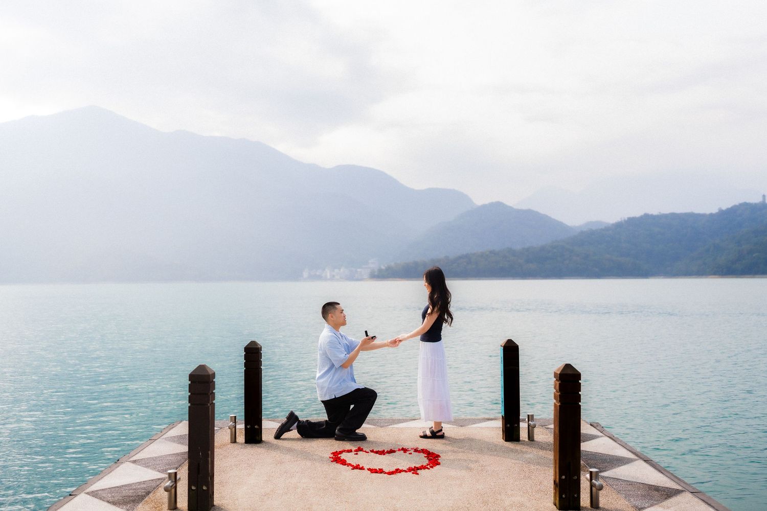 Man proposes to woman on a scenic lakeside pier with rose petals heart, mountains in background.
