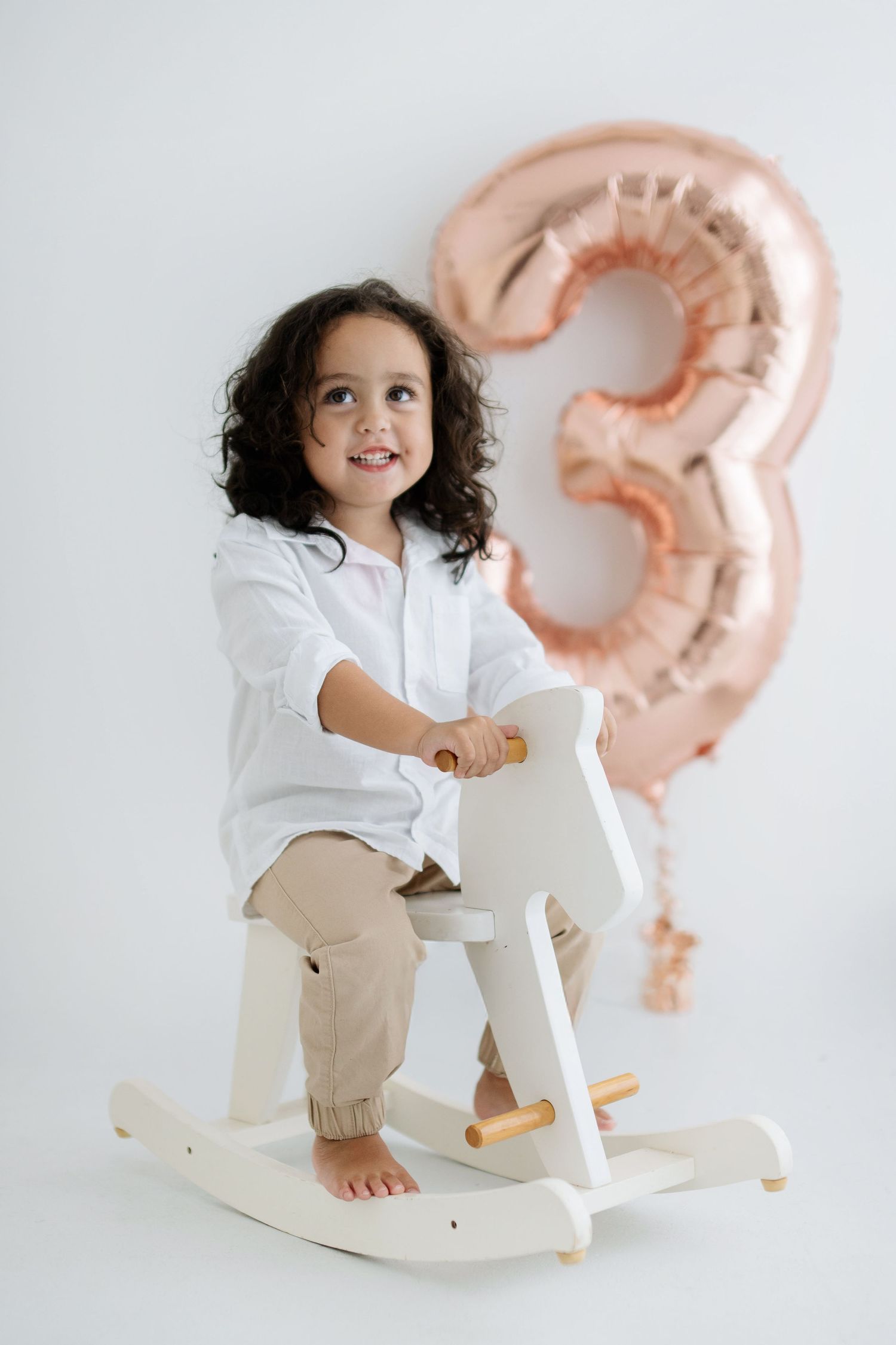 Smiling toddler girl riding a white wooden rocking horse beside a rose gold number 3 balloon on white background.