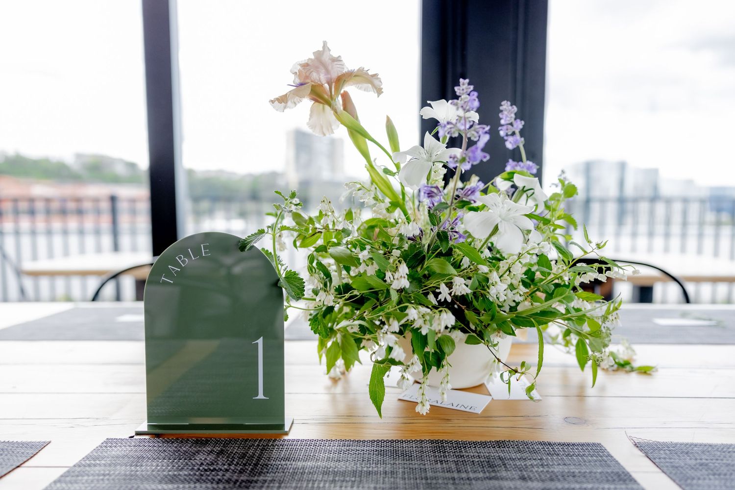 Bright rooftop restaurant dining room with floor-to-ceiling windows, black chairs, wooden tables, and city skyline views.