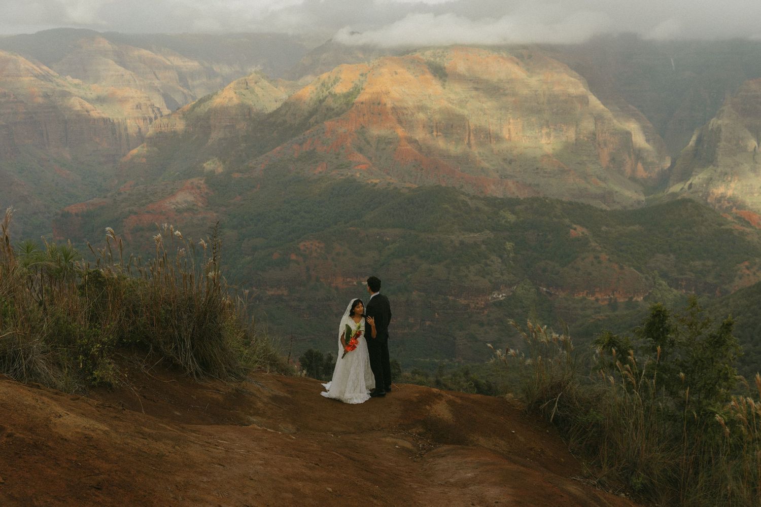 A couple stands silhouetted against dramatic mountain scenery at sunset.