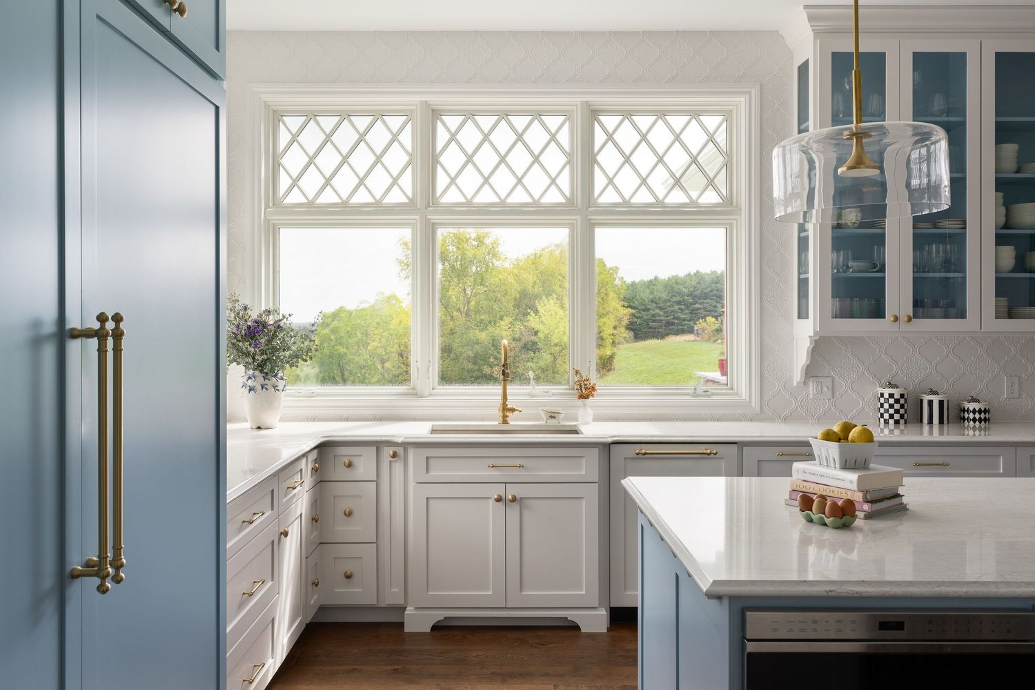Bright white kitchen with diamond-paned windows, marble countertops, and a blue accent island overlooking scenic outdoor views.