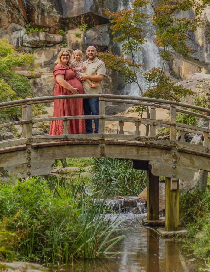 maternity session with mom dad and daughter embracing on a bridge with a waterfall in the background. Maternity photographer.