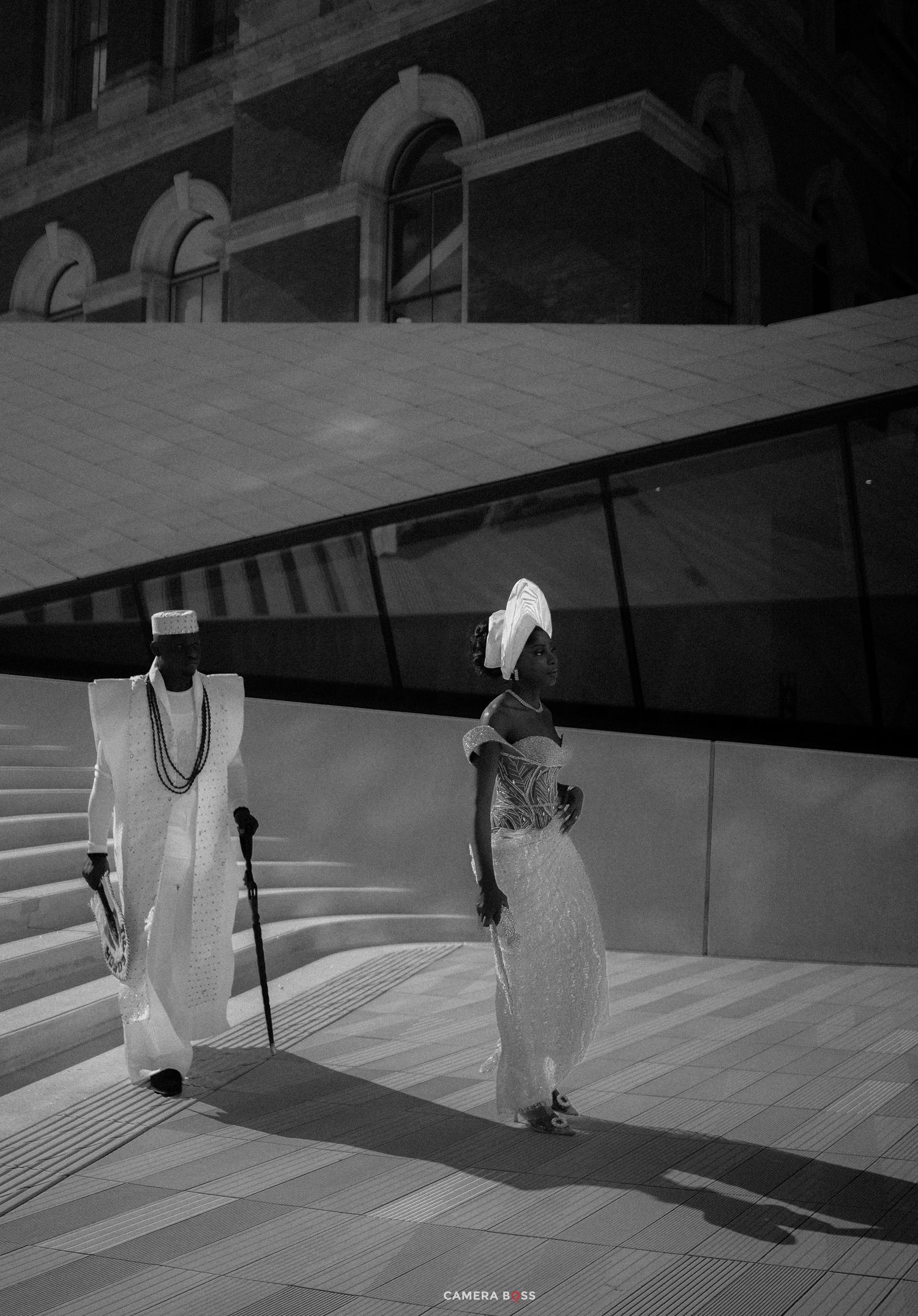 Two elegantly dressed individuals in white traditional attire walk near a modern building in a black and white photo.