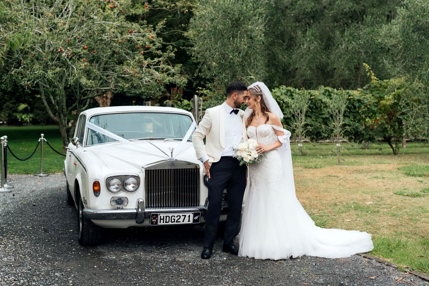 Bride and groom kissing beside a classic white Rolls-Royce adorned with white ribbons on a gravel driveway.