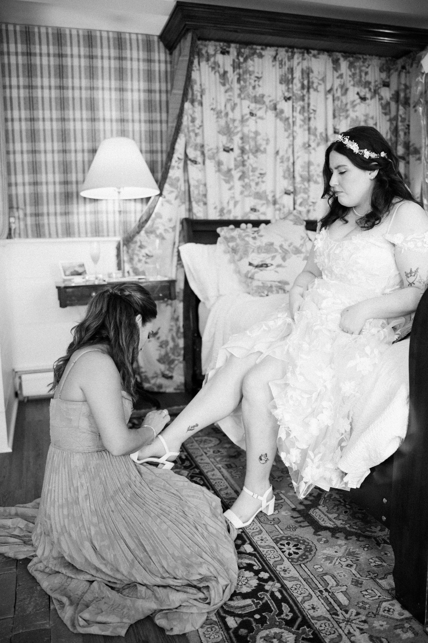 Bridesmaid helping bride put on wedding shoes in a hotel room, black and white bridal preparation photo.