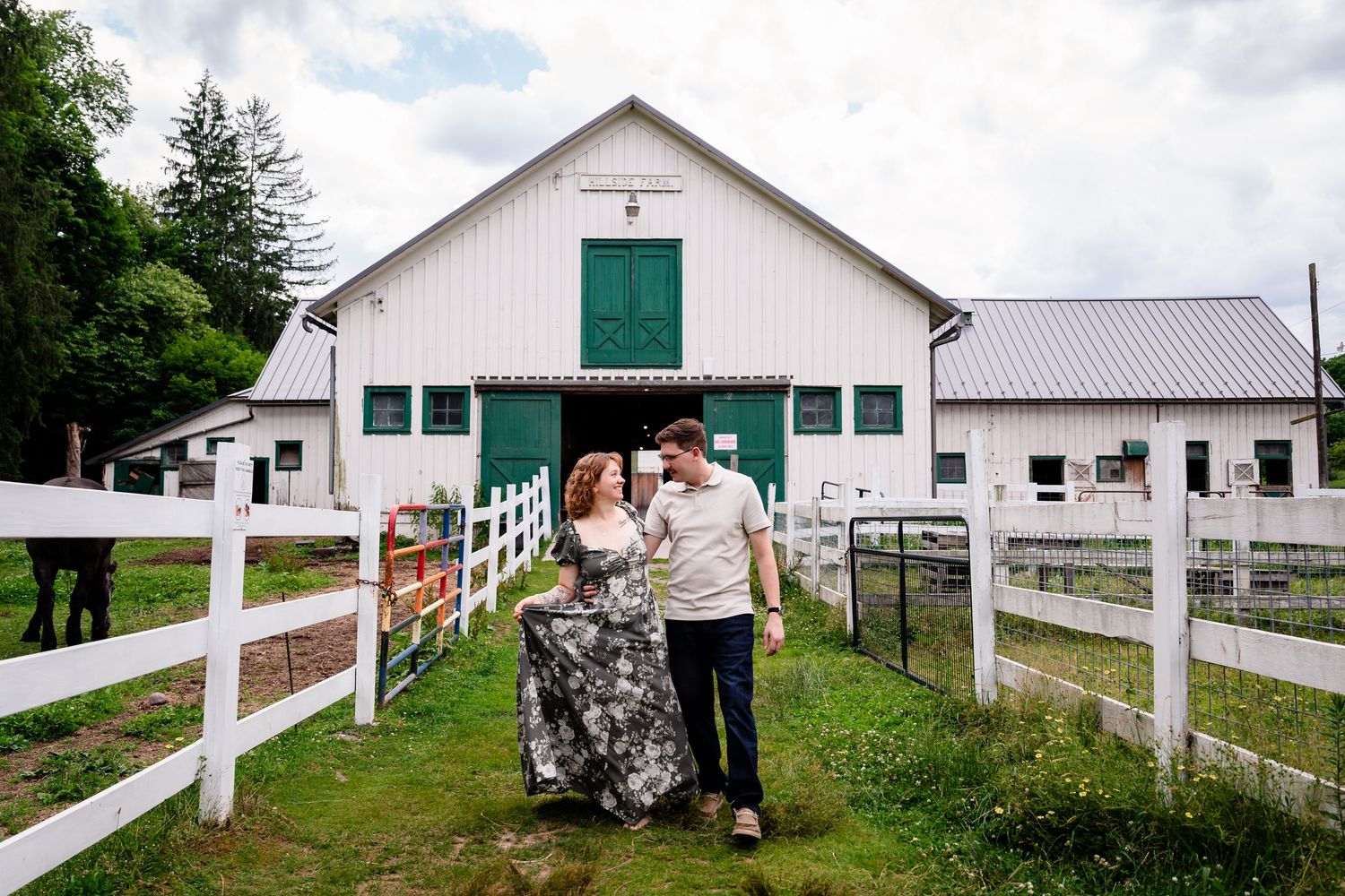 Couple walking hand in hand in front of a white barn with green doors on a lush farm property.
