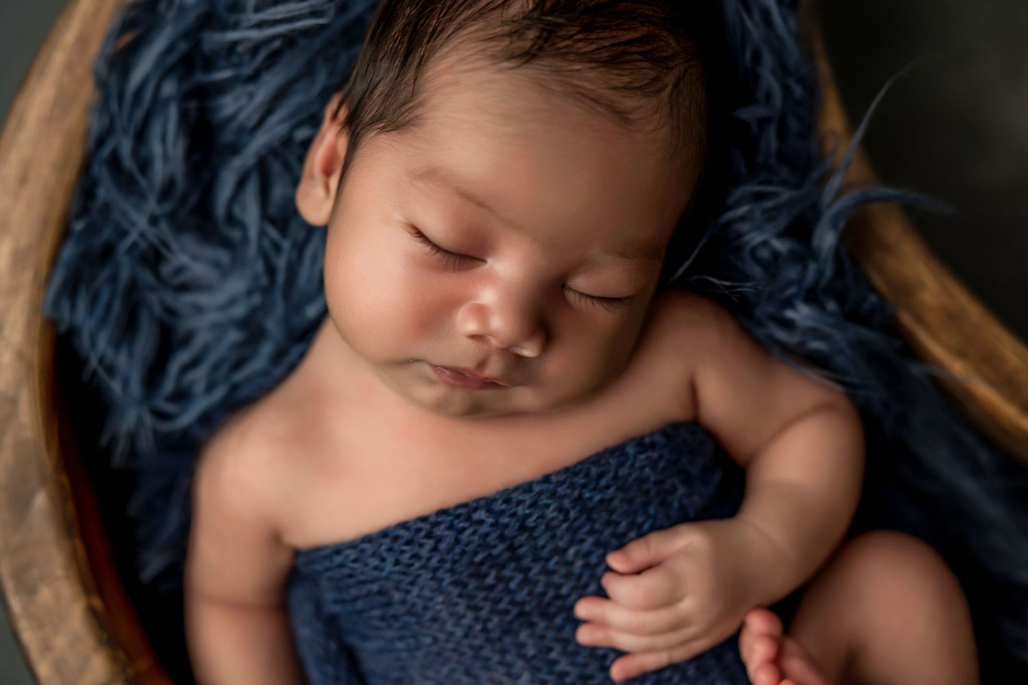 Sleeping newborn baby wrapped in a navy blue knit blanket, nestled in a wooden bowl with blue fluffy layers.
