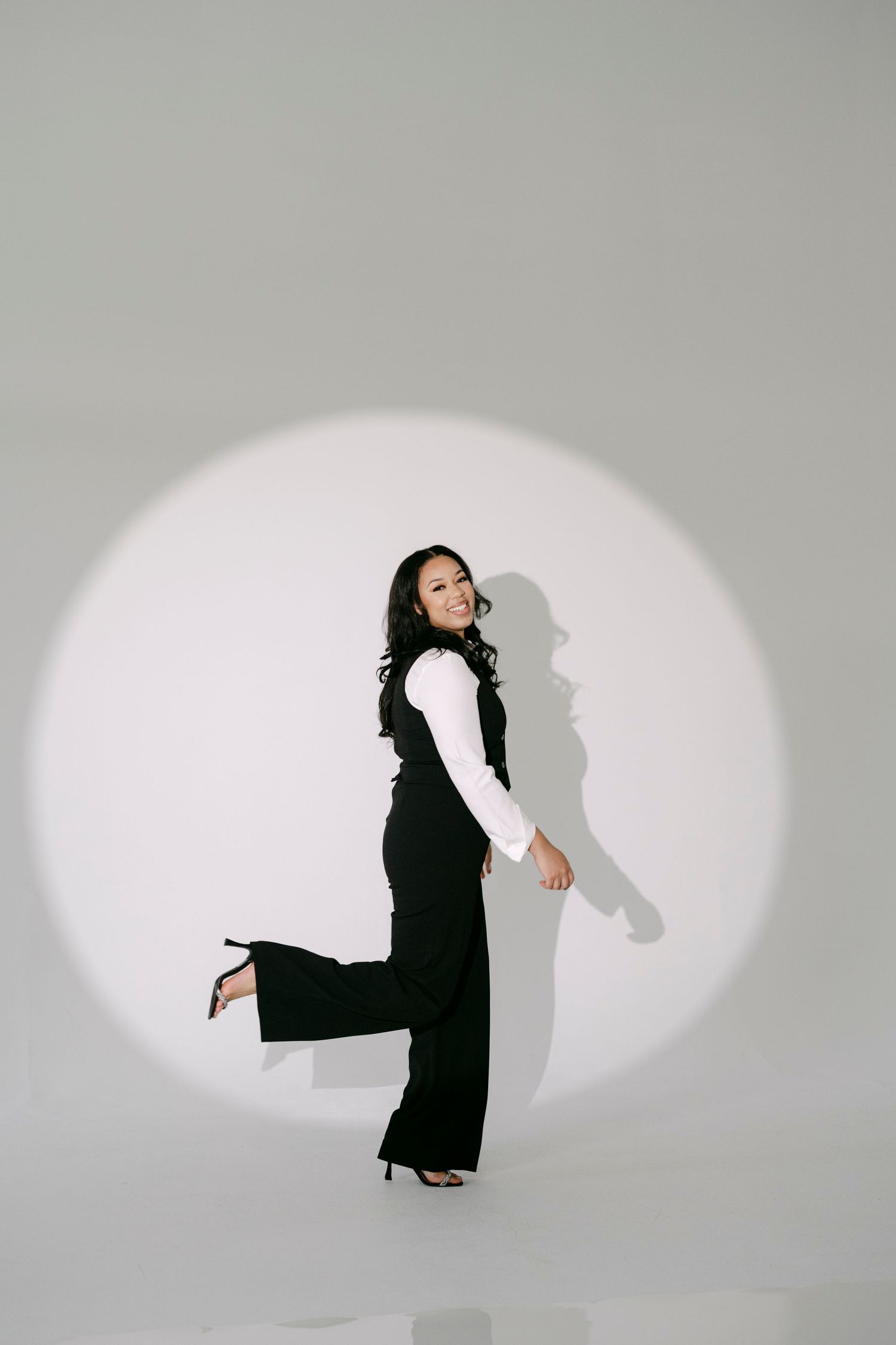 A woman in black wide-leg pants and vest poses confidently in a spotlight against a white background.