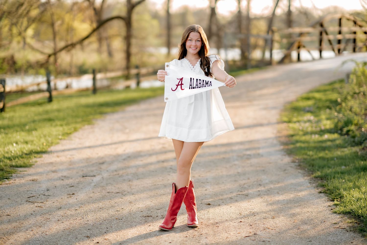 Young woman in white Alabama dress and red cowboy boots walks a tree-lined path holding an Alabama pennant.