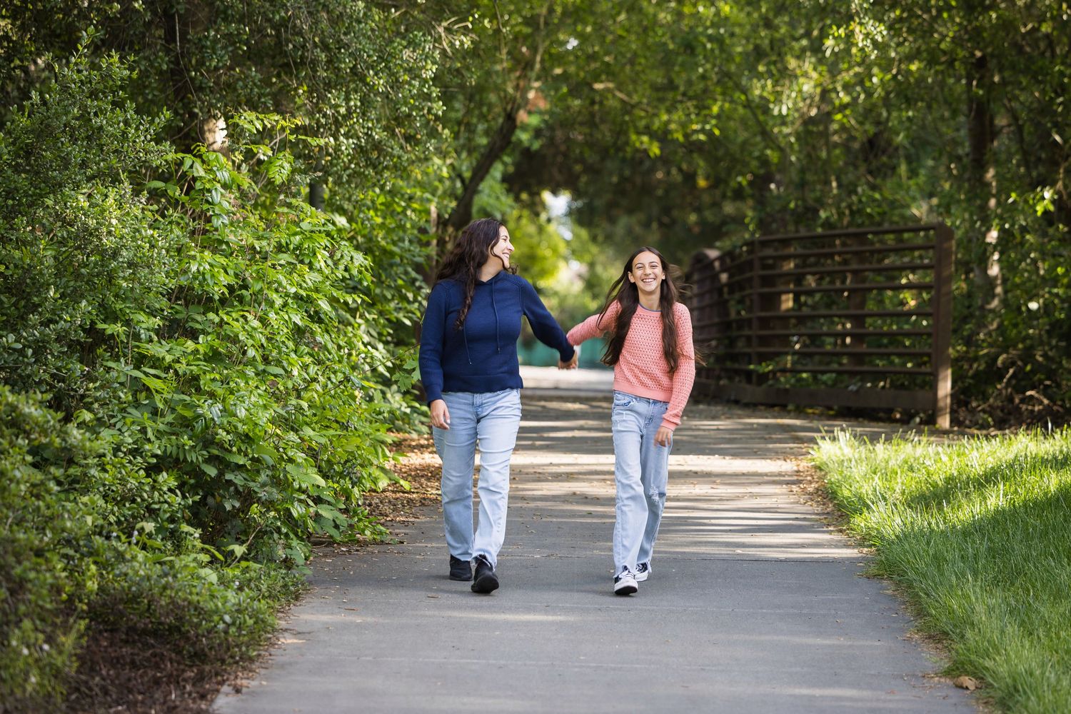 Two women holding hands and walking together on a sunny park path surrounded by lush green trees.