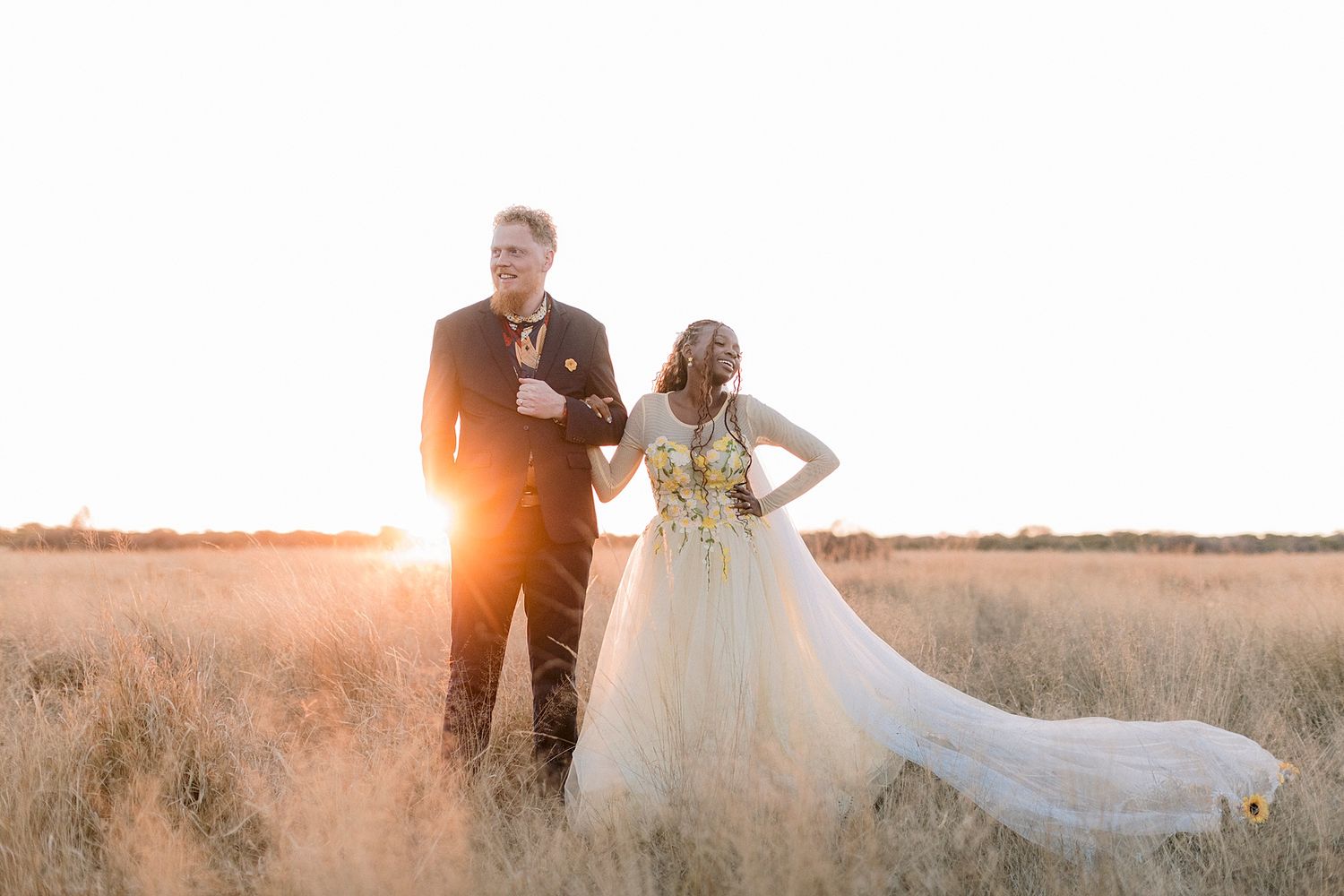 Couple poses in golden field at sunset, bride in flowing white gown with gold details, groom in black tuxedo.
