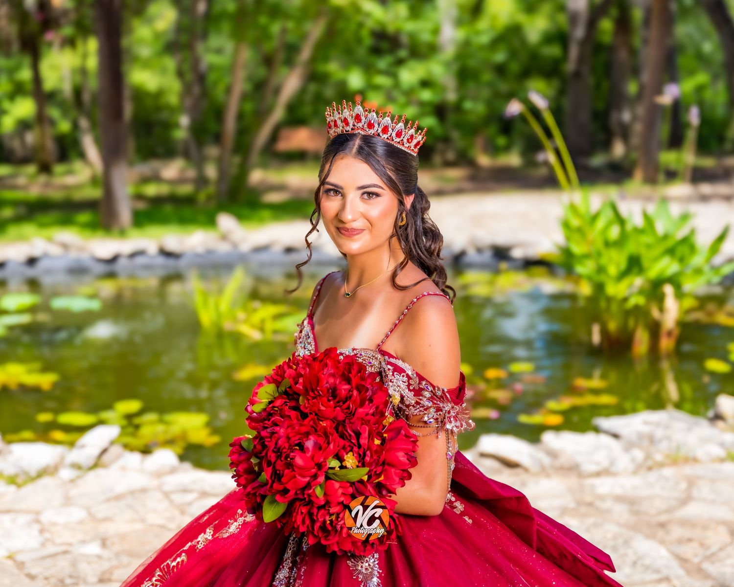 Young woman in red quinceañera gown holding red roses bouquet, wearing crown, posing near pond in lush garden.