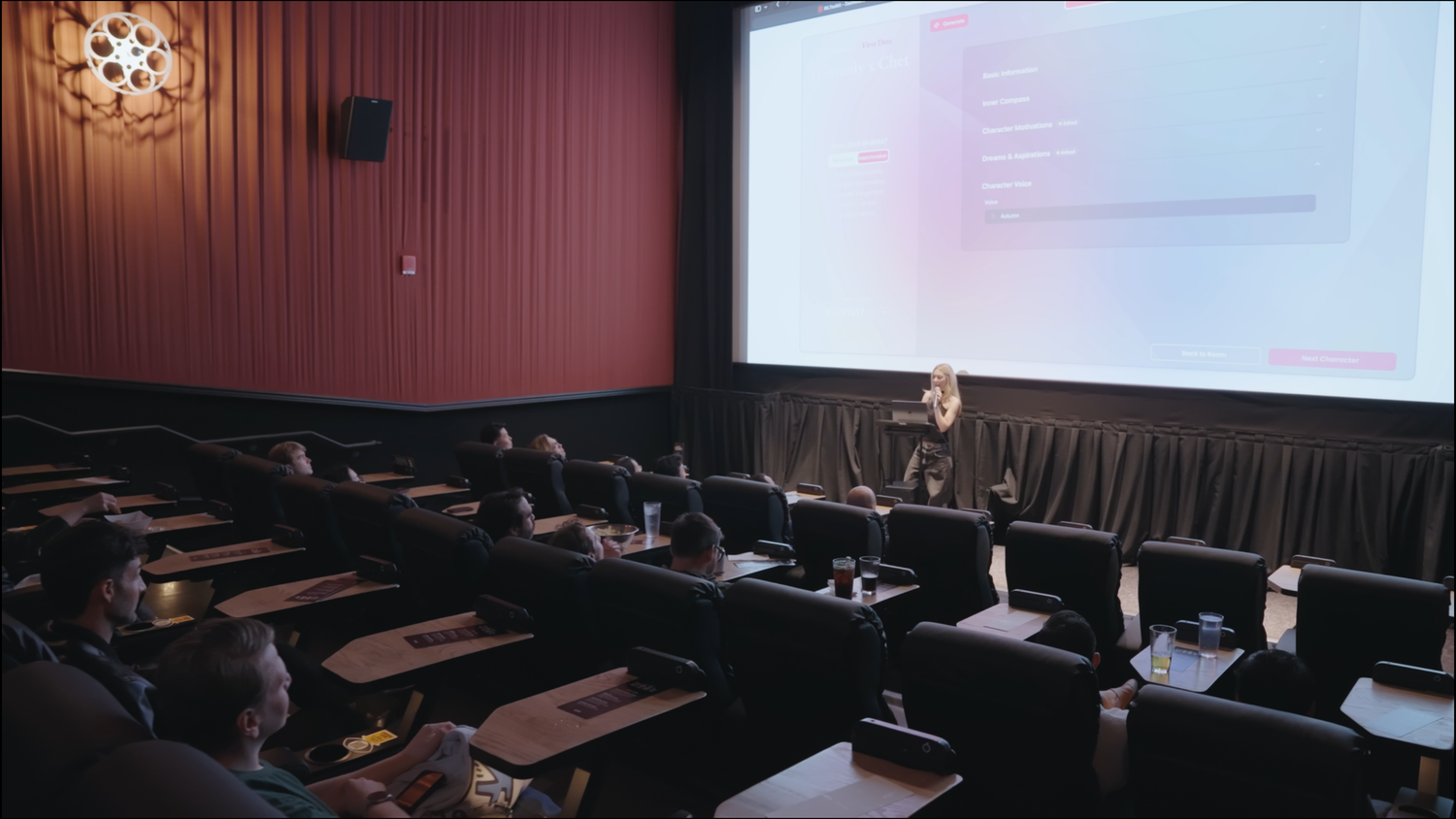 A presenter speaks at a podium in a cinema-style conference room with tiered seating and a large projection screen.