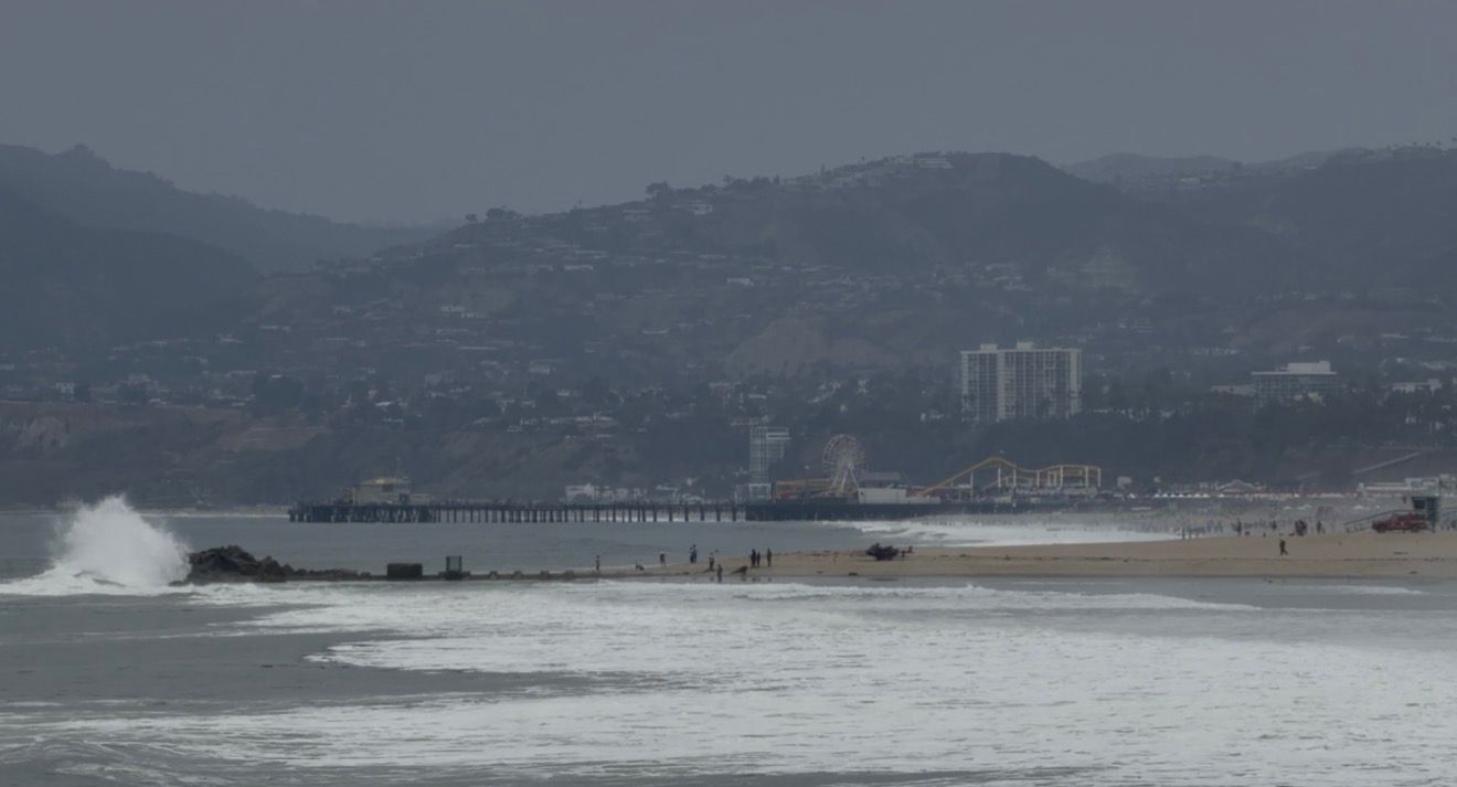 Waves crash near a pier on a hazy beach with hills and city buildings in the background.