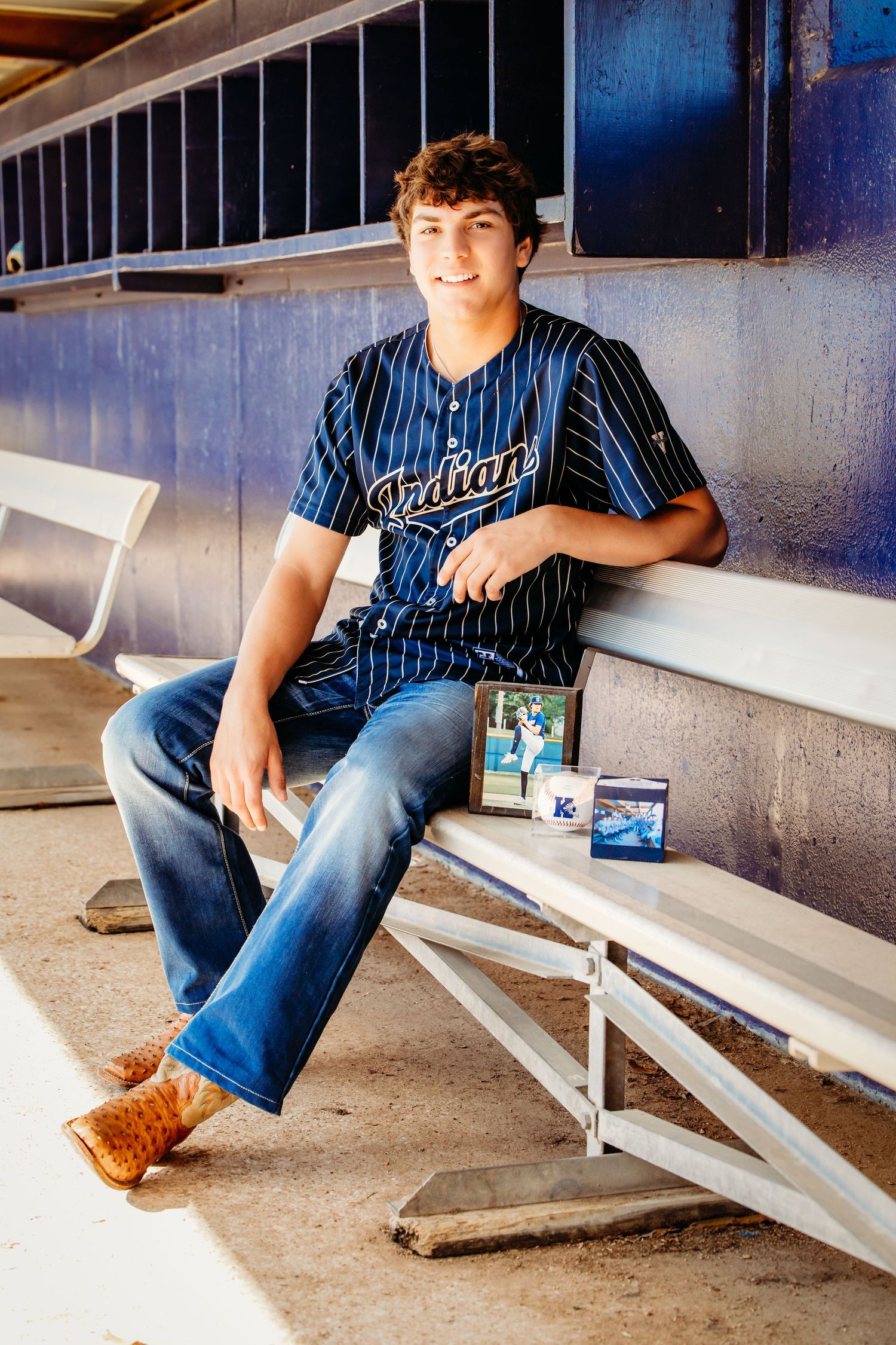 Teen boy in Indians baseball jersey sits in dugout during Keller, TX senior portrait session