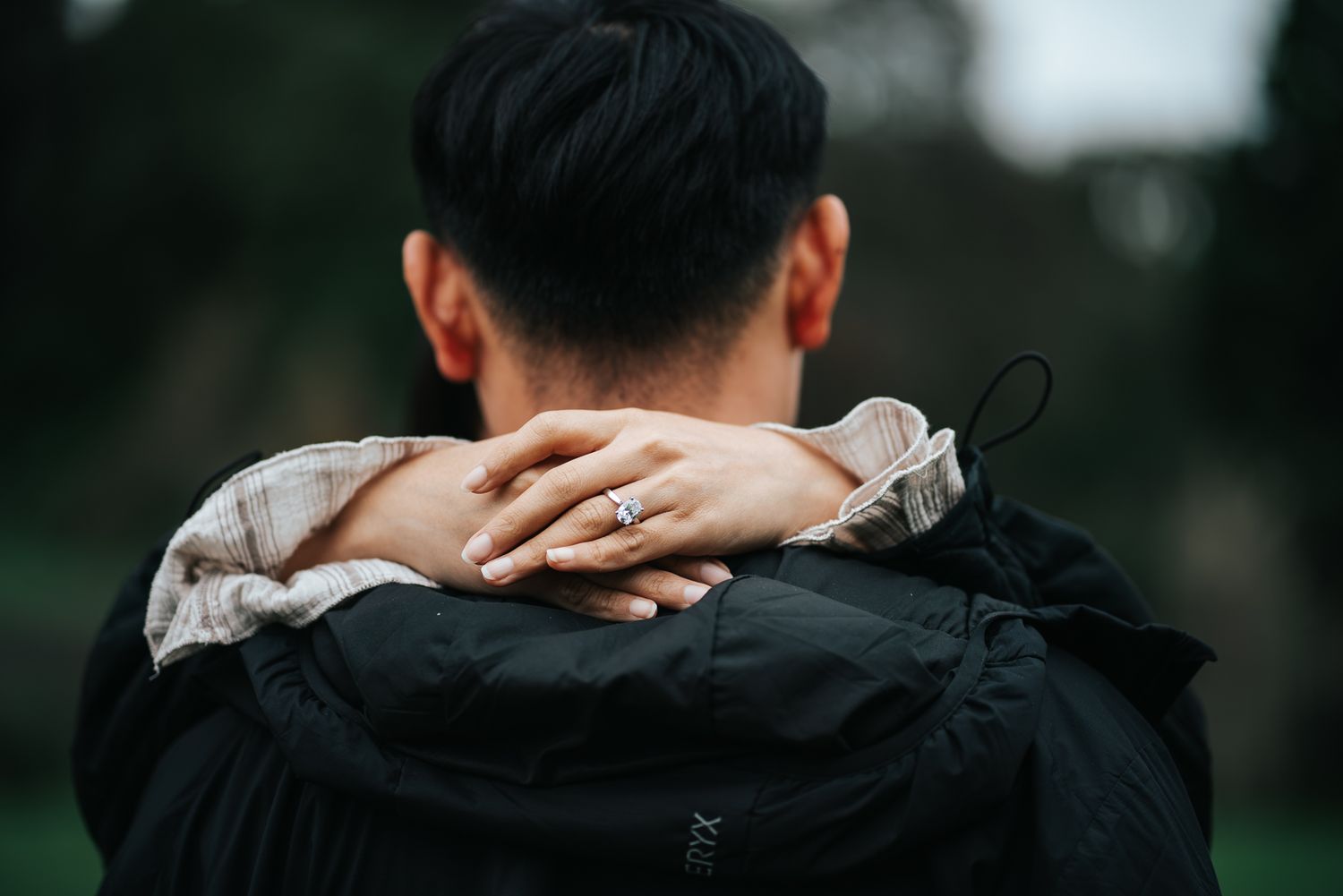 Woman with engagement ring wraps arms around mans neck from behind in moody outdoor setting taken by Melbourne Proposal Photographer.