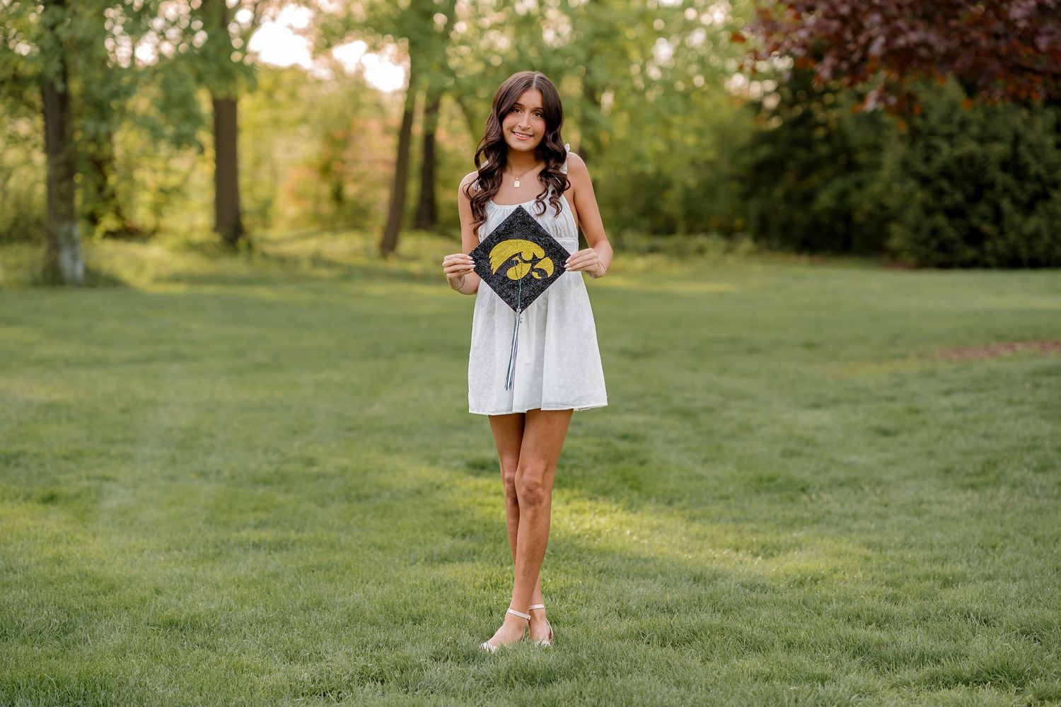 Young woman in white dress holds graduation cap on green lawn, celebrating her University of Iowa graduation.