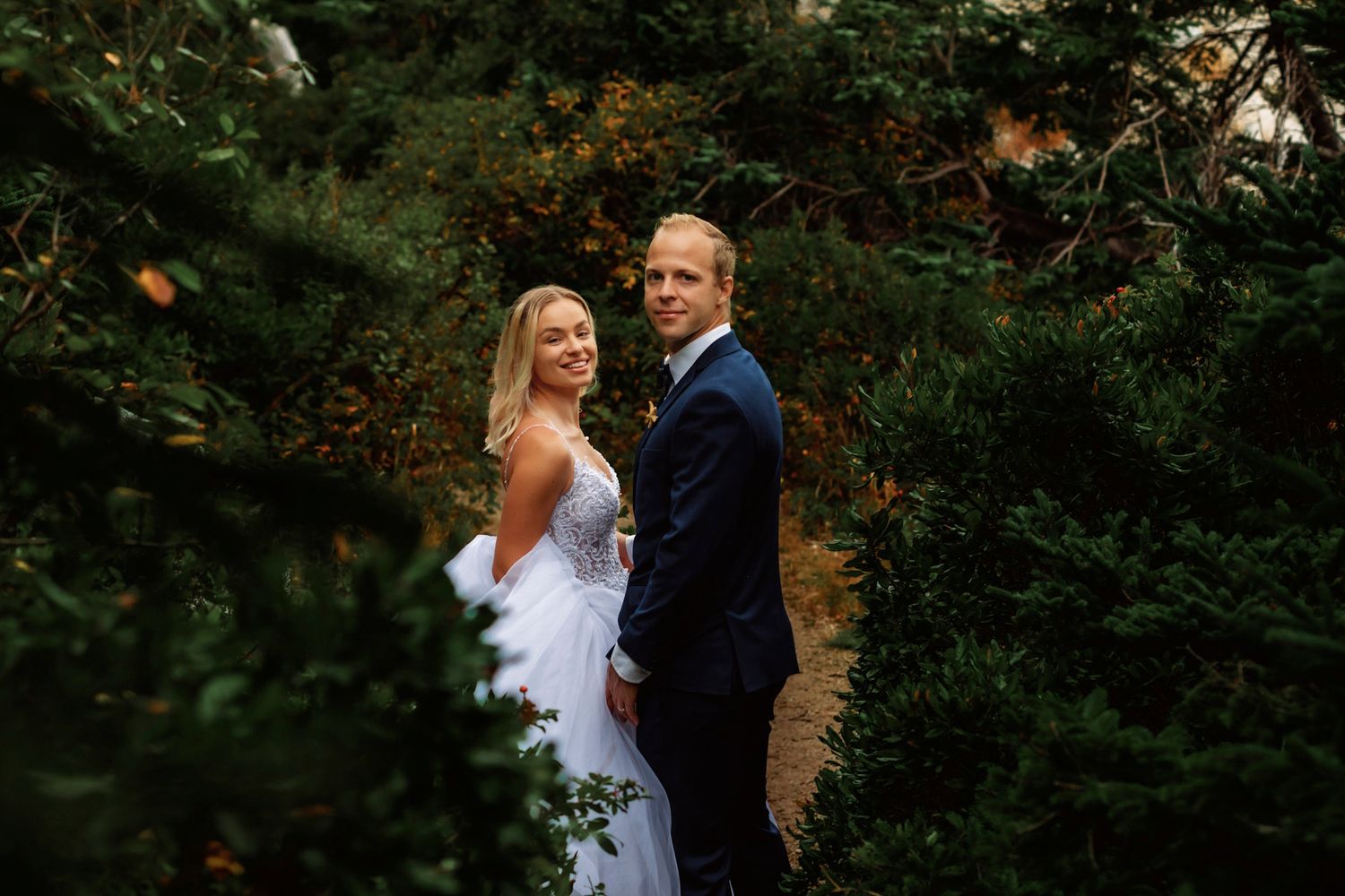 Bride and groom posing together among lush green trees in a romantic outdoor wedding portrait.
