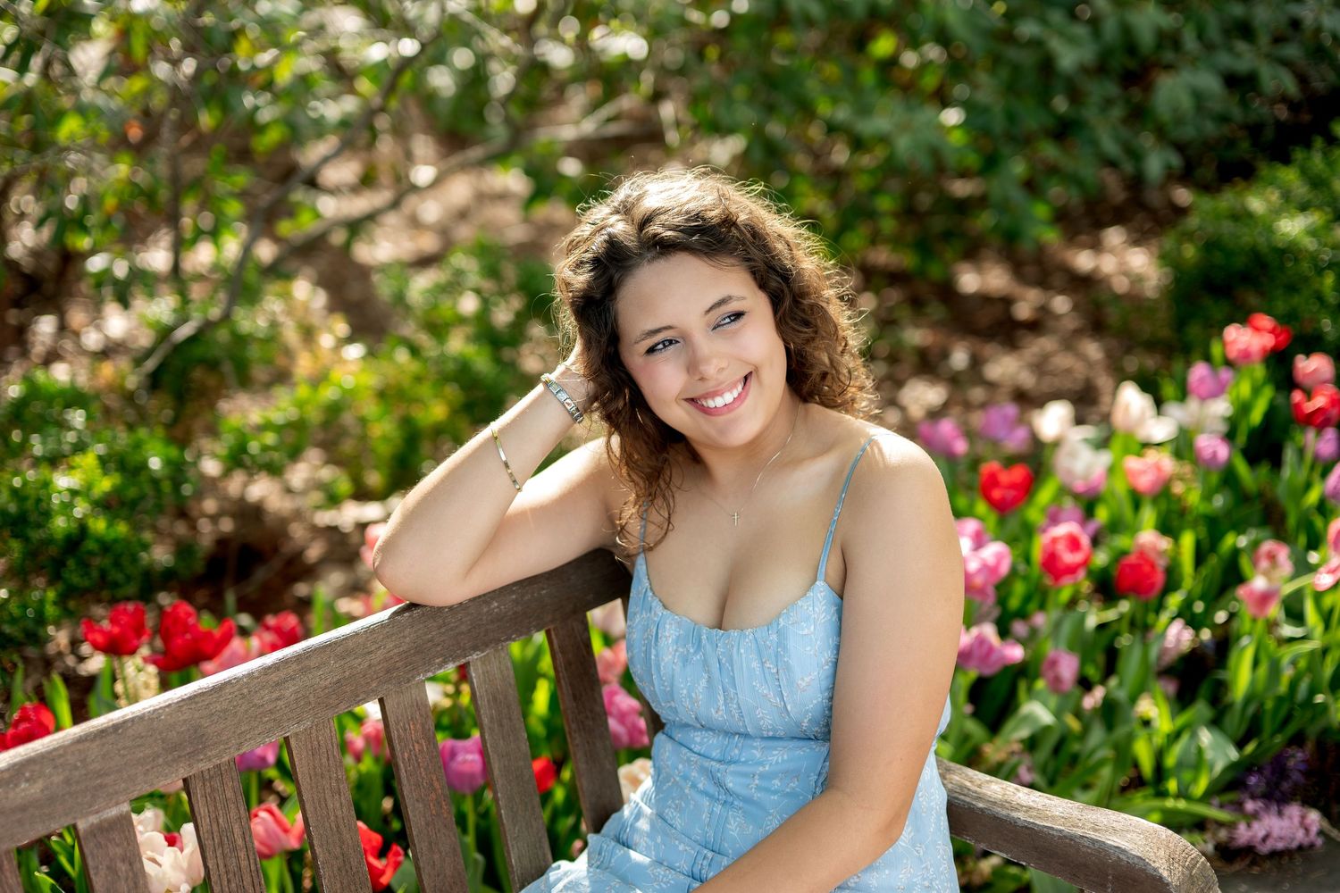 Young woman with curly hair smiling on a wooden bench surrounded by colorful tulips in a garden.