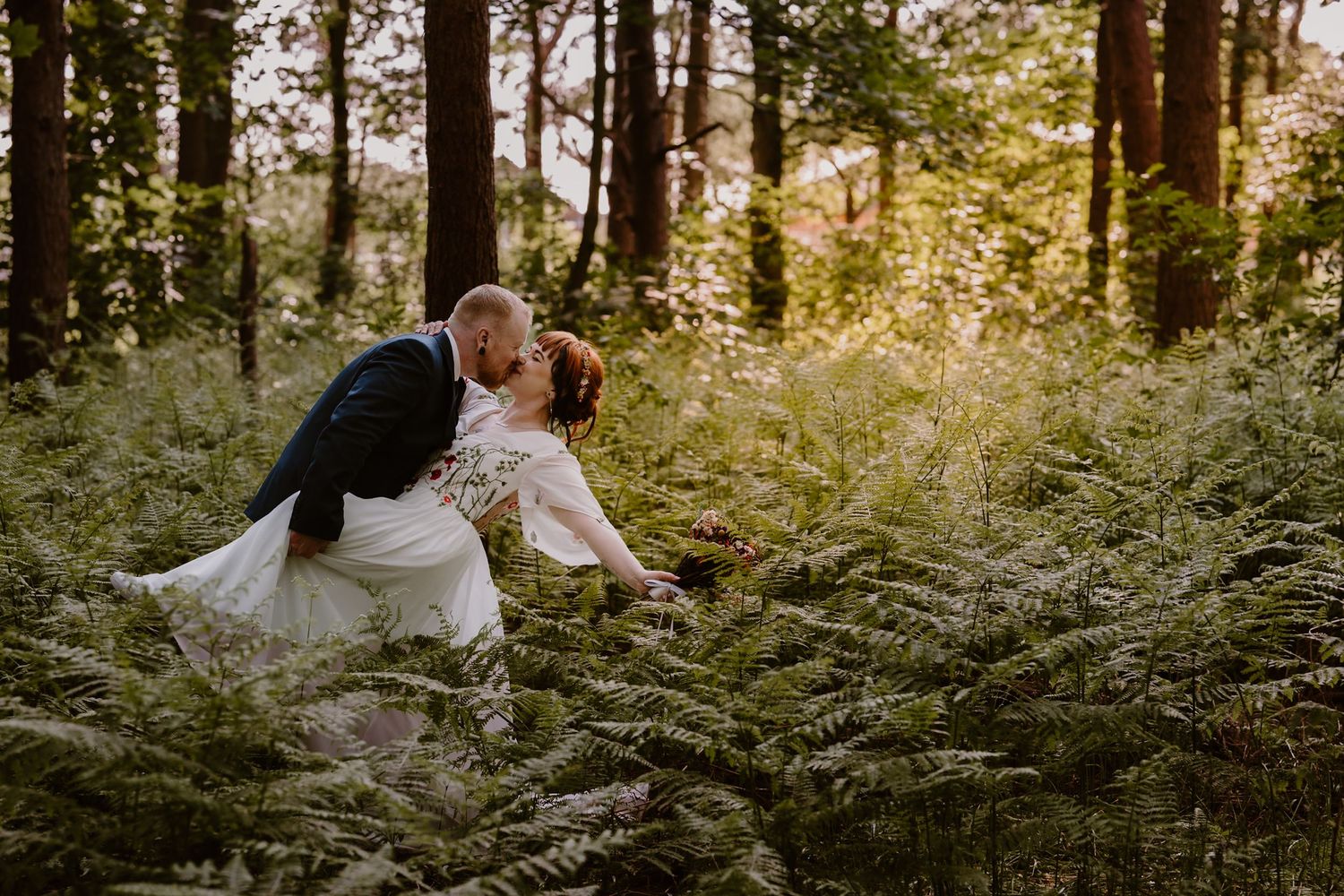Bride and groom share a kiss outdoors as confetti falls around them, bride in white strapless gown, groom in blue tweed suit.