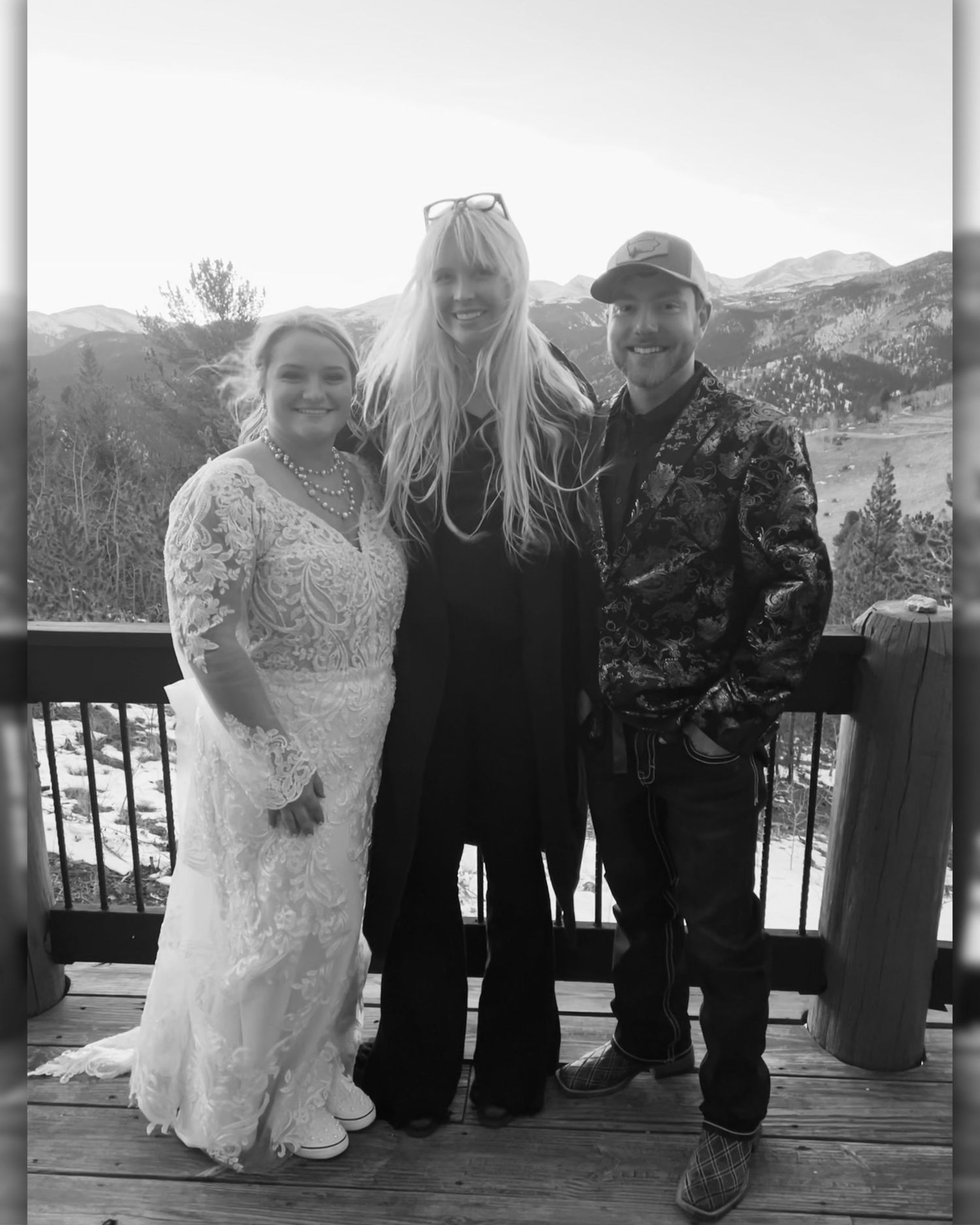 Black and white photo of a bride in lace gown, woman in black, and man in camo jacket posing on a mountain deck.