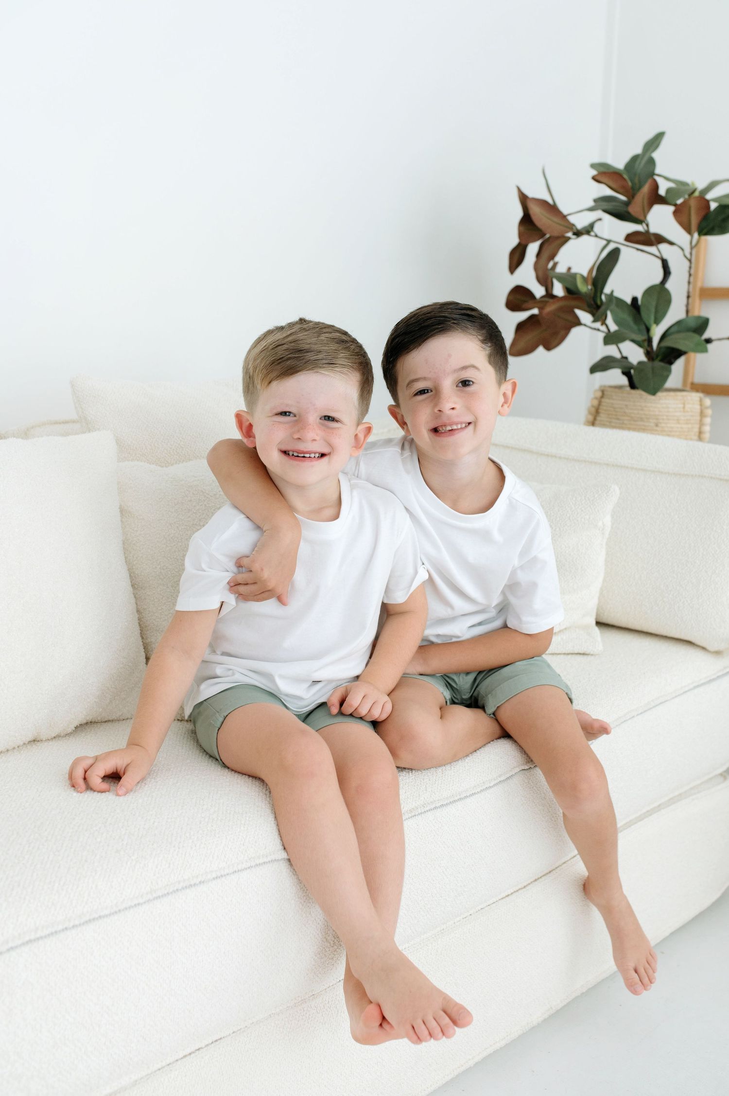 Two smiling young boys in white t-shirts and green shorts hugging on a white couch indoors.