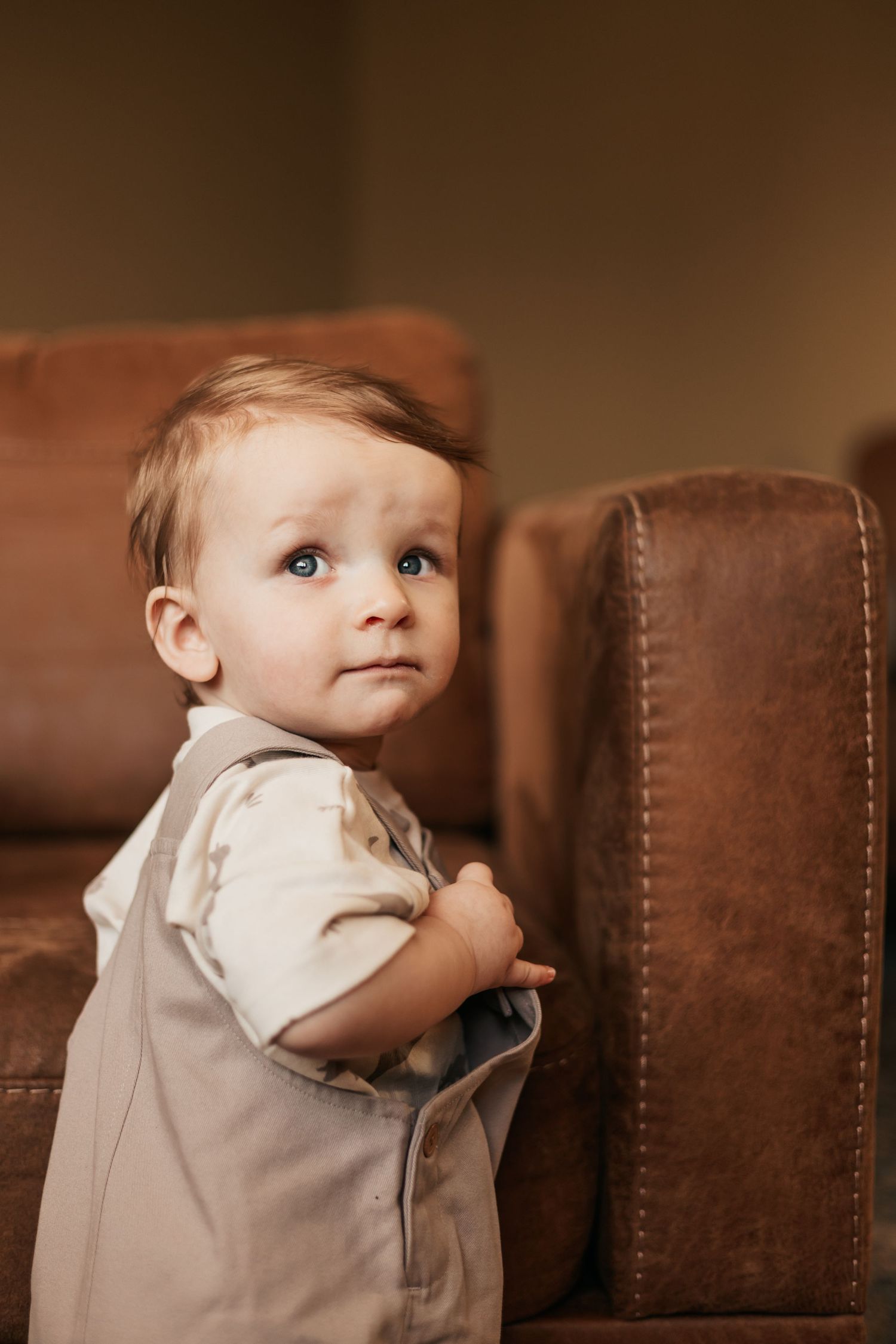Adorable baby boy in suspenders standing next to a brown leather sofa, looking up | Professional Milestone Sessions for baby