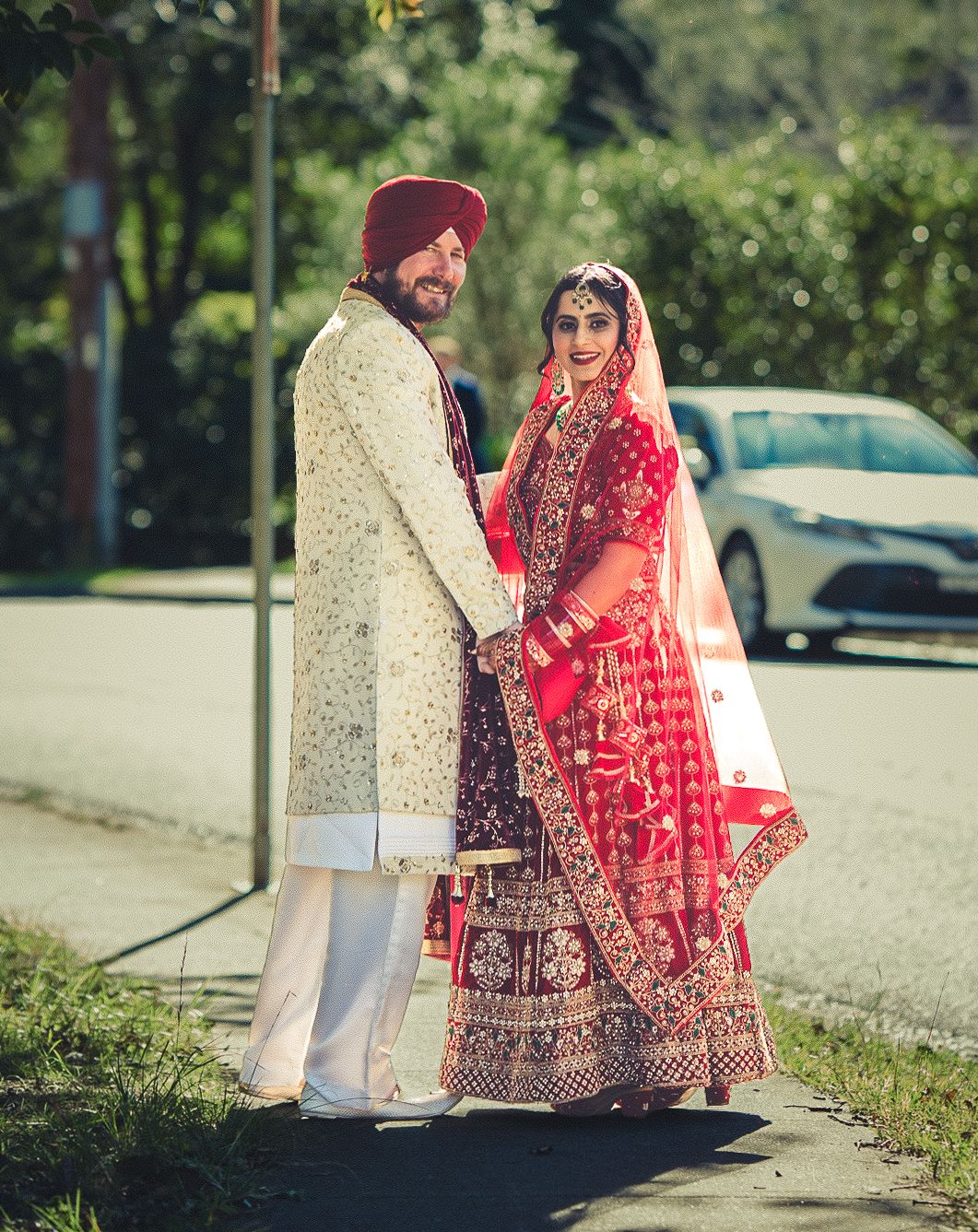 Sikh couple in traditional wedding attire, groom in white sherwani and red turban, bride in red lehenga with dupatta.