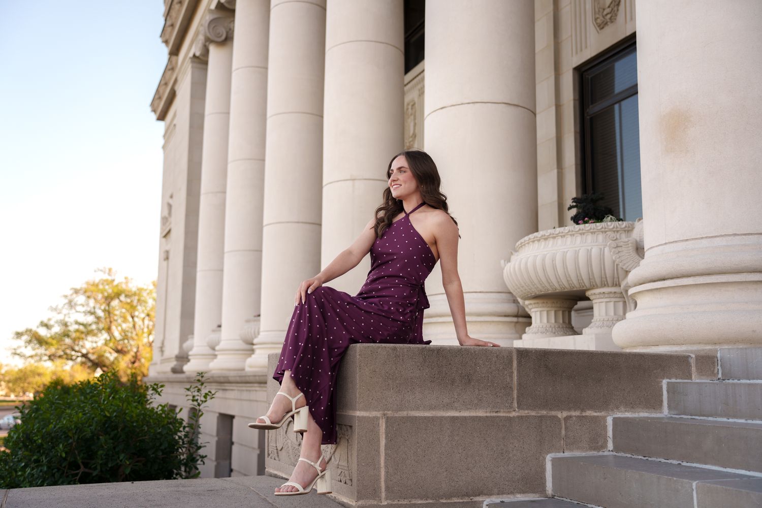 Young woman in burgundy floral dress poses on stone steps of elegant columned building at sunset.