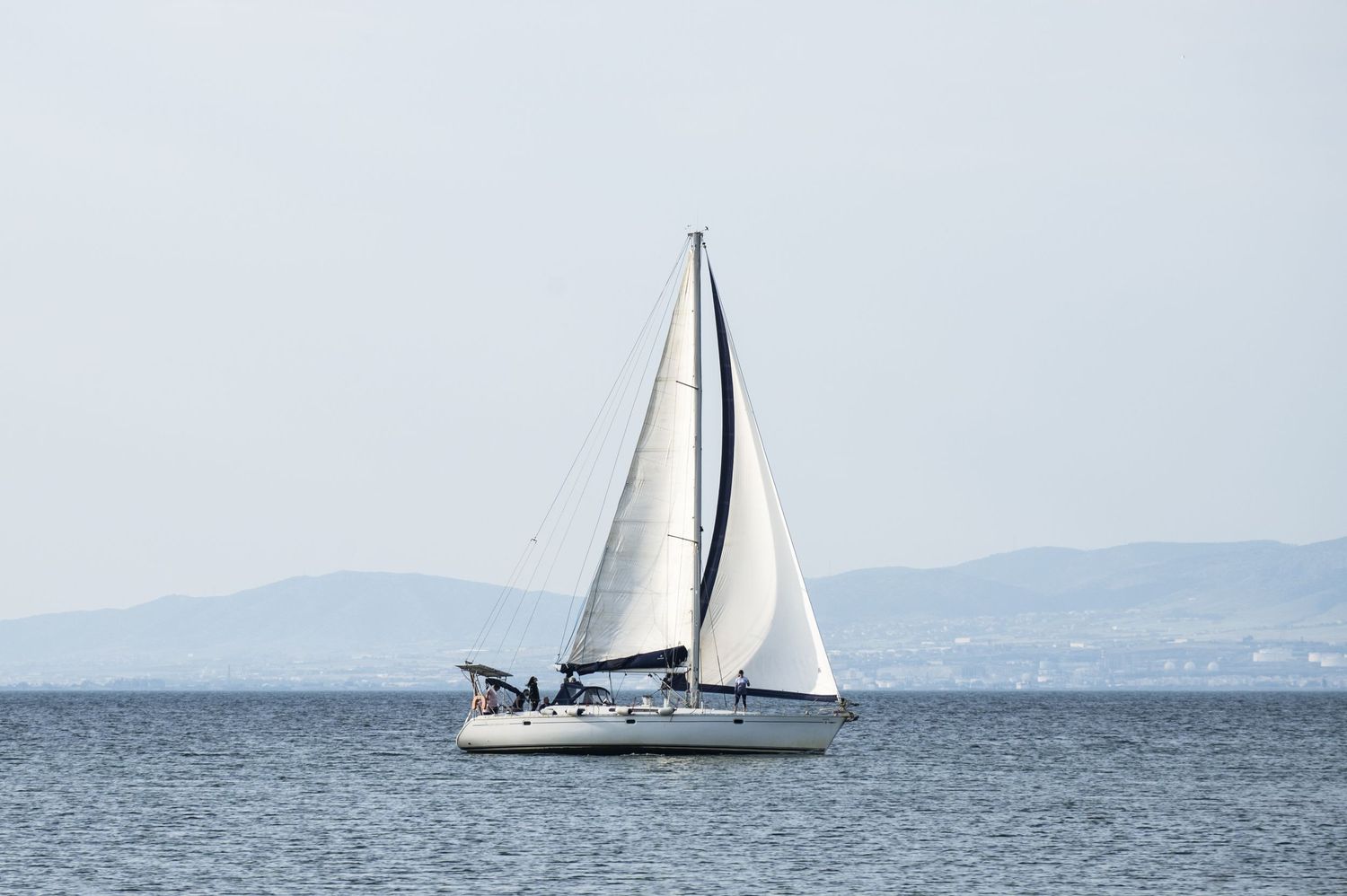 A white sailboat glides across calm blue waters with distant mountains visible on the horizon.