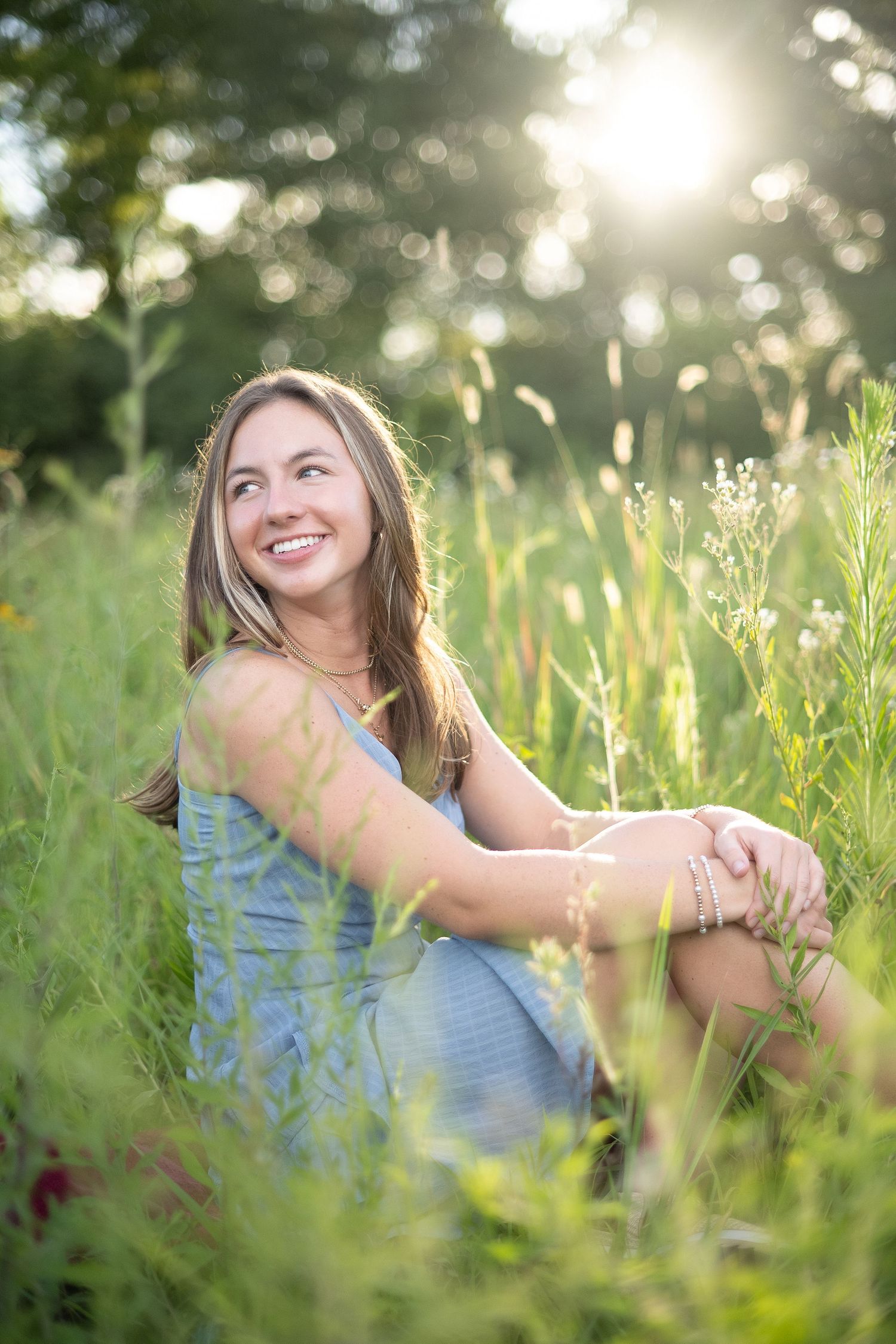 Young woman smiling in a wheat field wearing a blue dress, with lush green trees in the background.