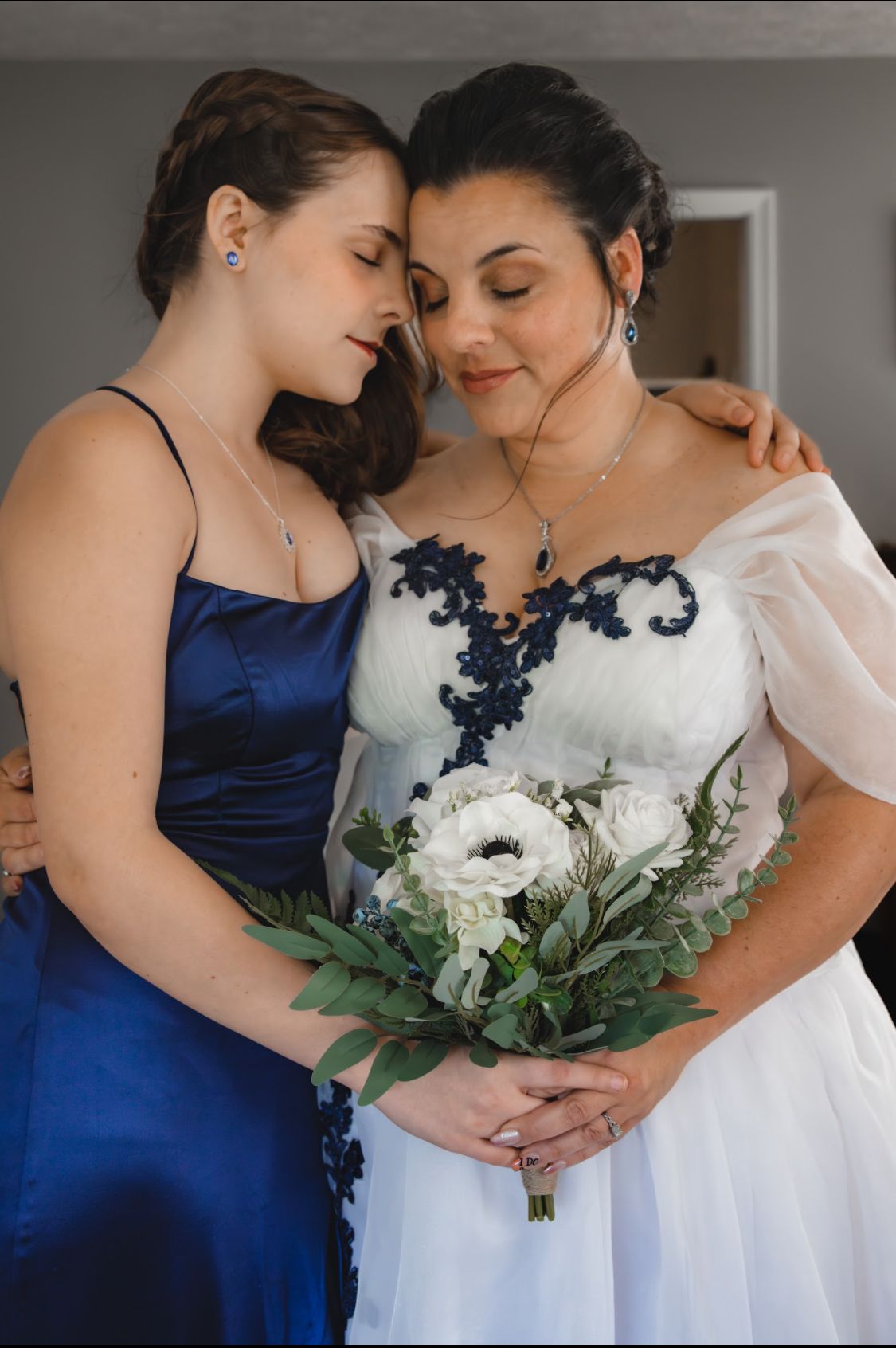 Bride in white gown with navy lace and bridesmaid in blue satin dress share tender moment holding white floral bouquet.