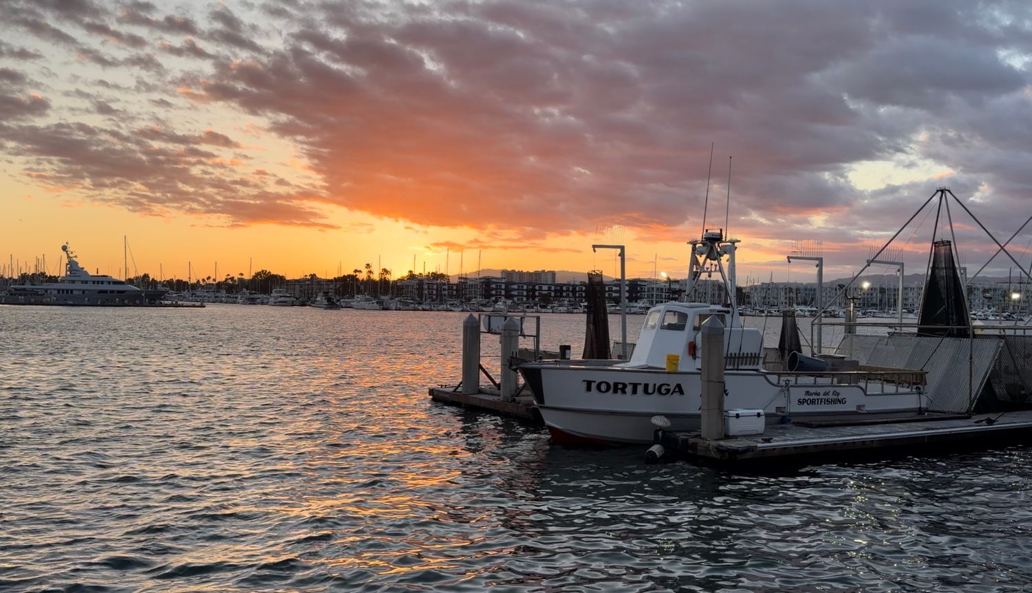 Boat named Tortuga docked at marina pier during vibrant orange and pink sunset over calm harbor waters.