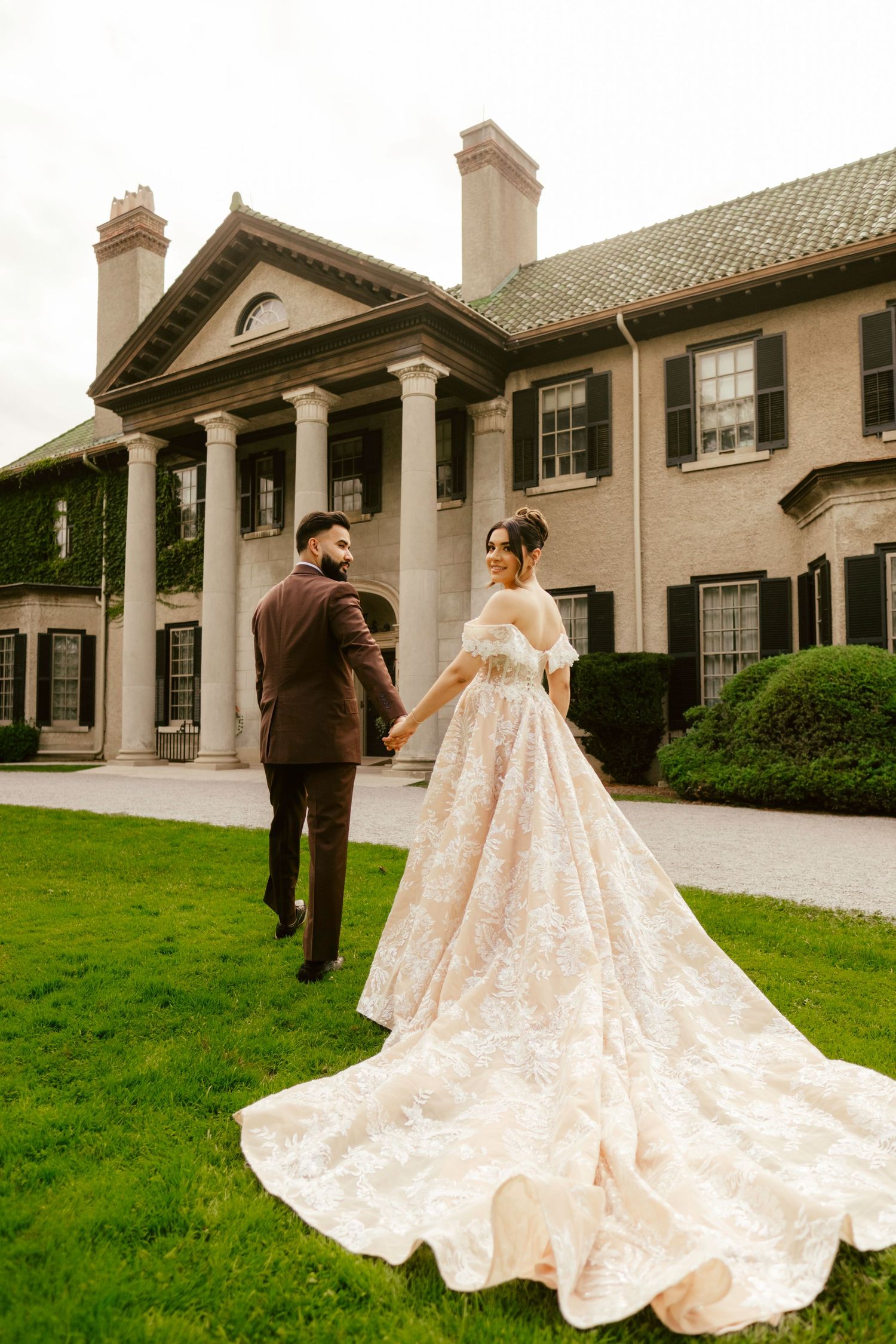 A couple in formal wedding attire stands on a manicured lawn in front of a grand mansion with classical columns.
