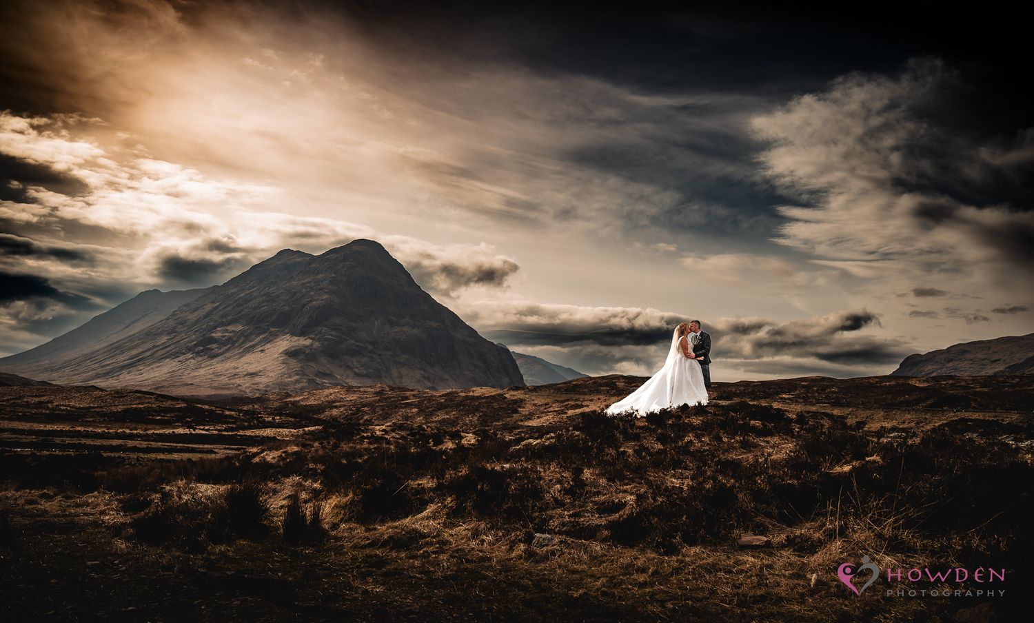 Wedding couple stands in dramatic mountain landscape under moody clouds during sunset.