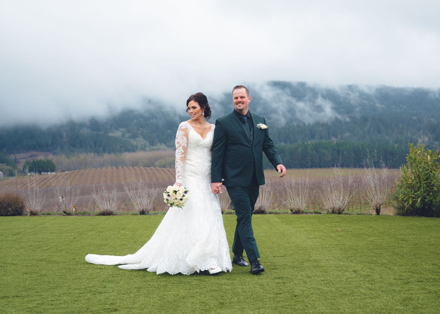 A couple stands on lush grass in their wedding attire with misty mountains and vineyards in the background.