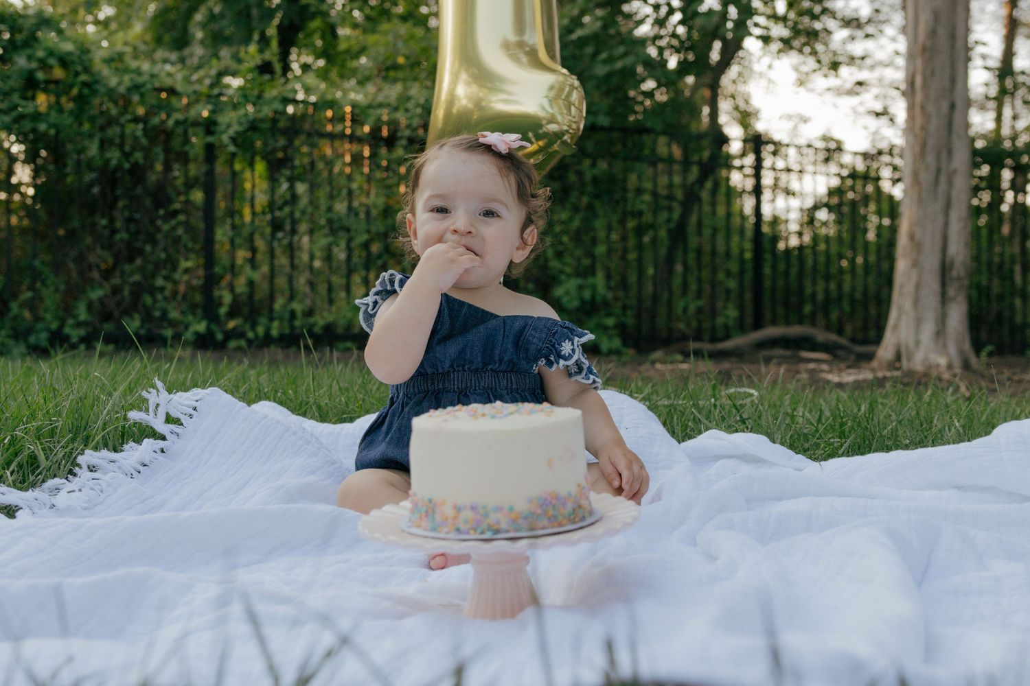 Baby in navy dress sitting on white blanket with gold balloon.