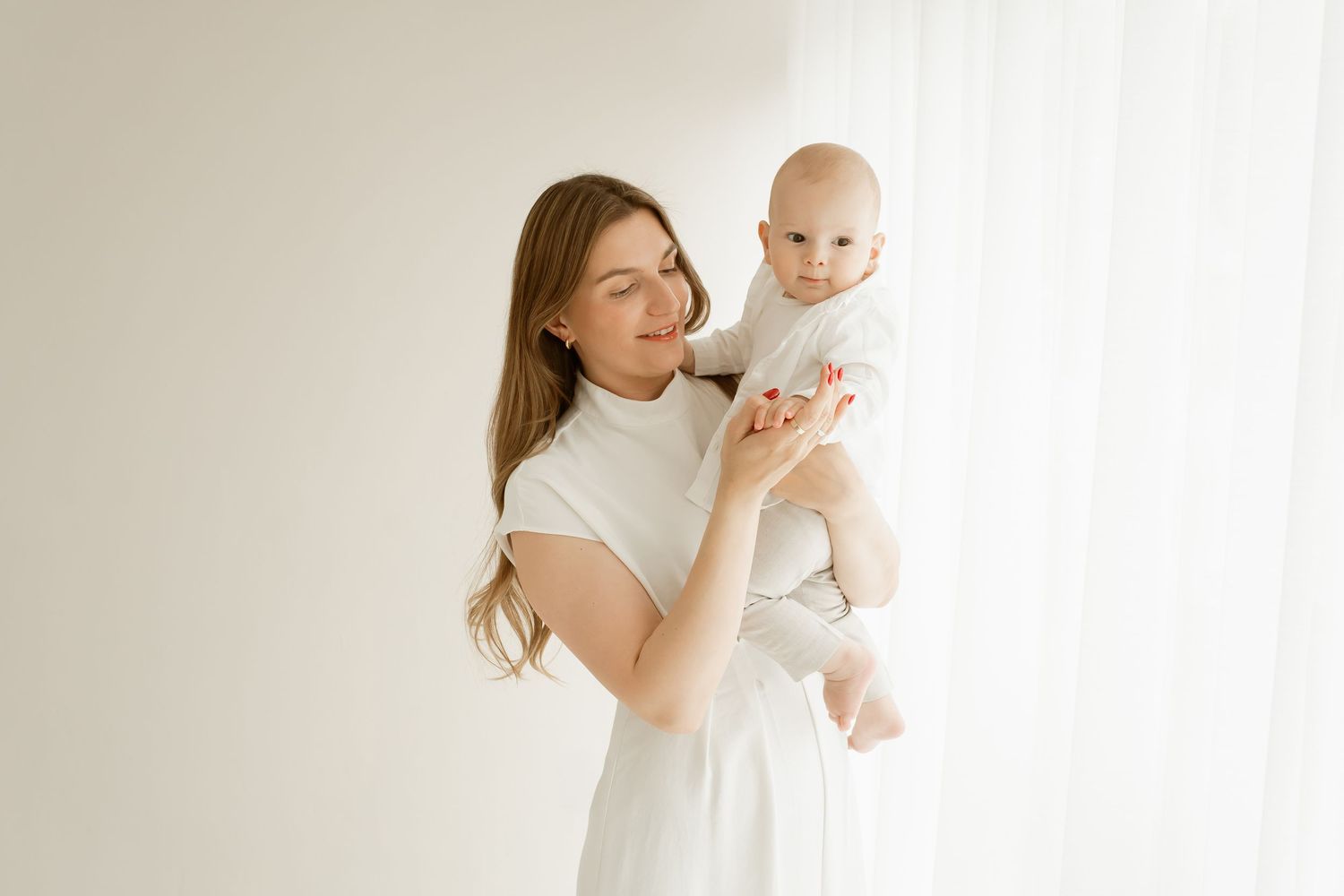 A parent in a white dress holds a smiling baby wearing light colored clothing against a bright minimal background.