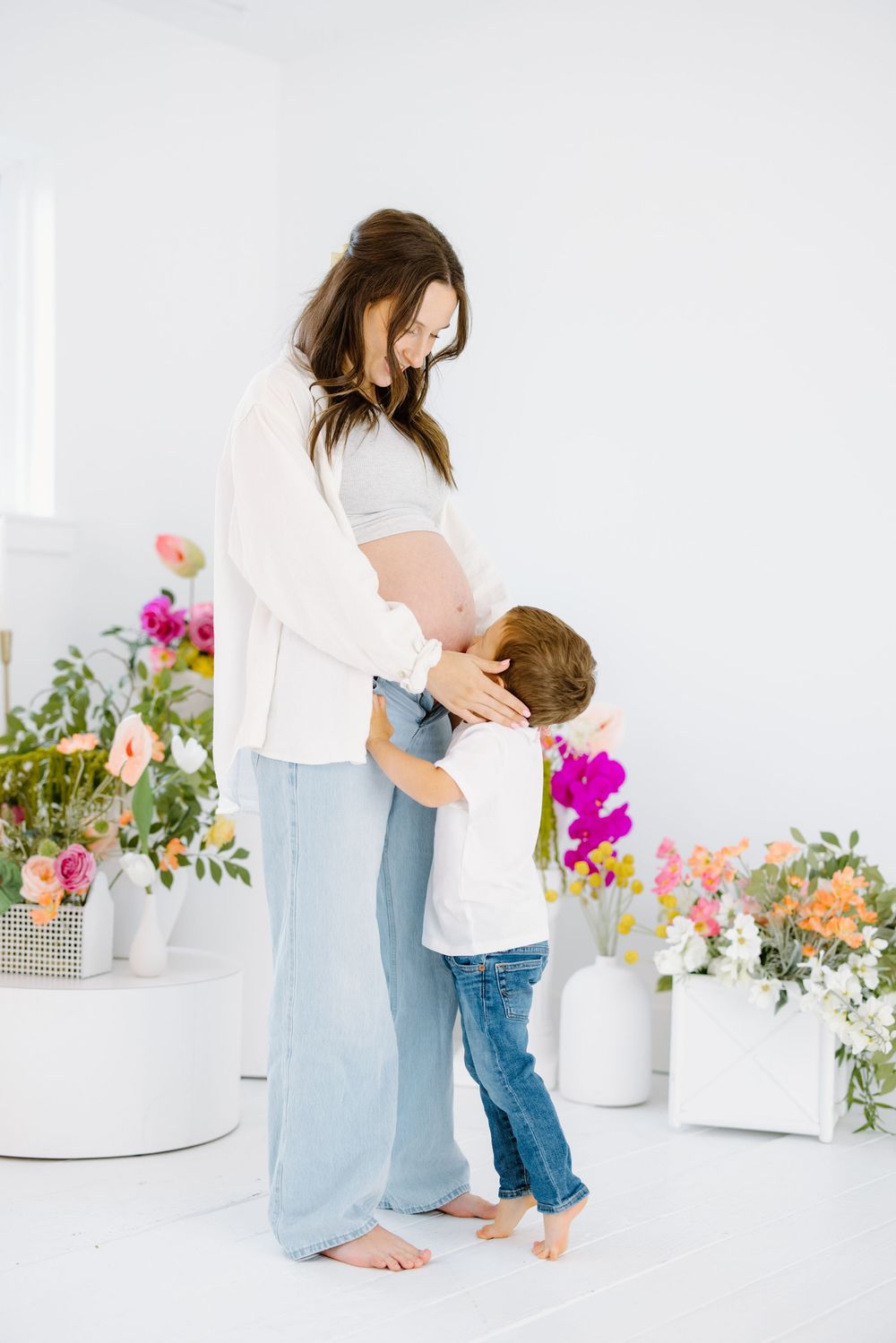A pregnant woman in white and denim embraces a young child in a bright room decorated with colorful flowers.