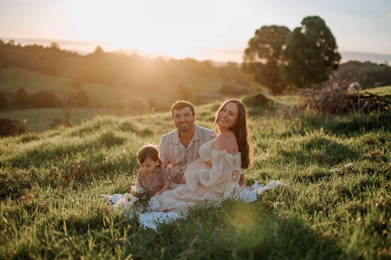 Family sitting together on blanket in grassy field during golden sunset with lens flare and trees in background.