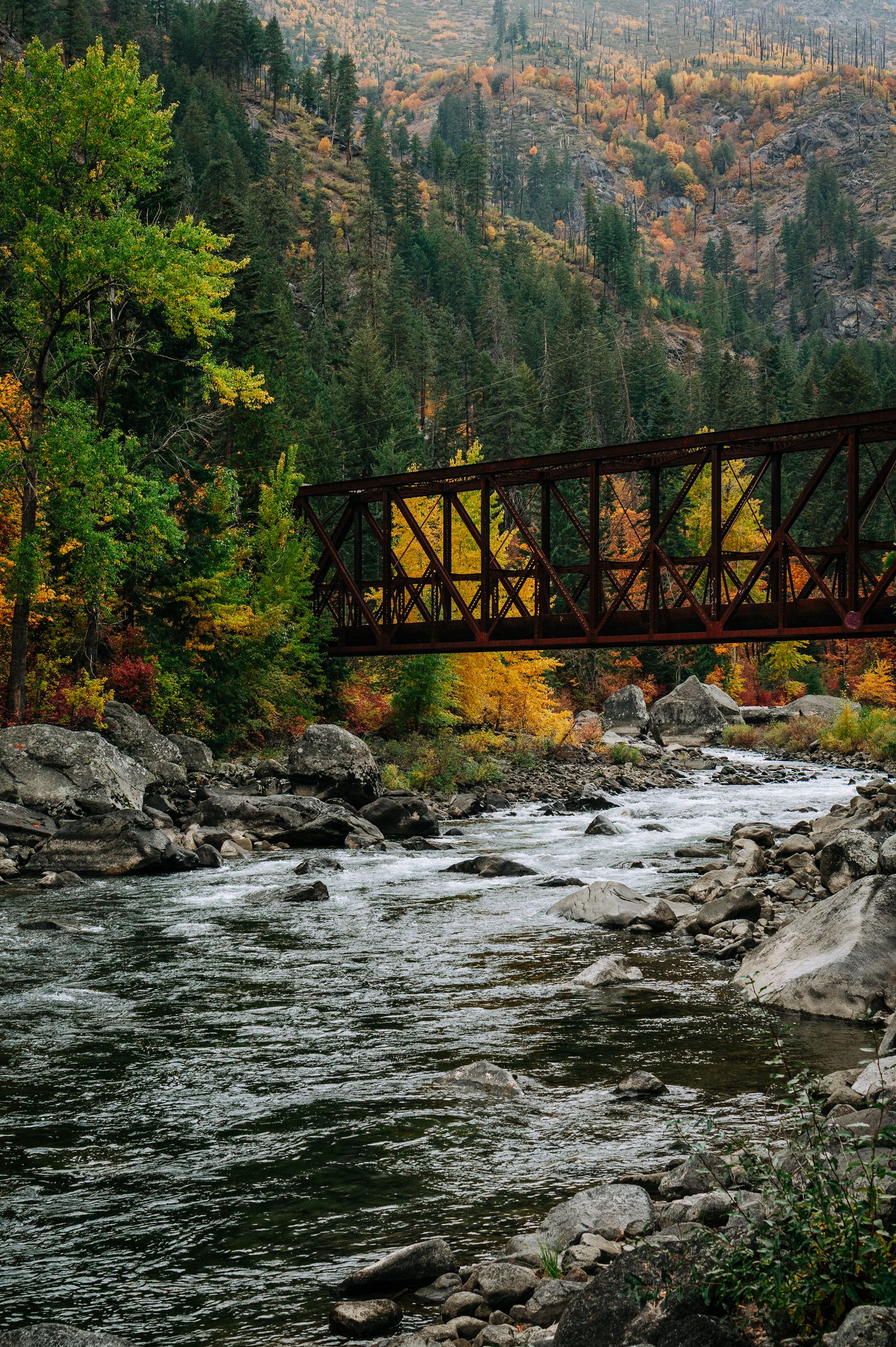 A rustic metal bridge spans across rushing river rapids surrounded by autumn foliage and rocky cliffs in a mountain setting.