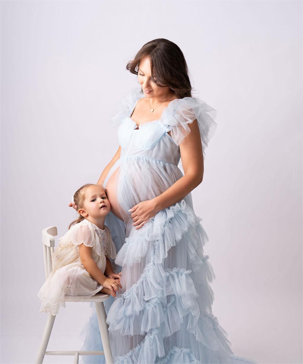 A tender maternity portrait showing a pale blue ruffled gown and small matching dress, set against a light studio backdrop.