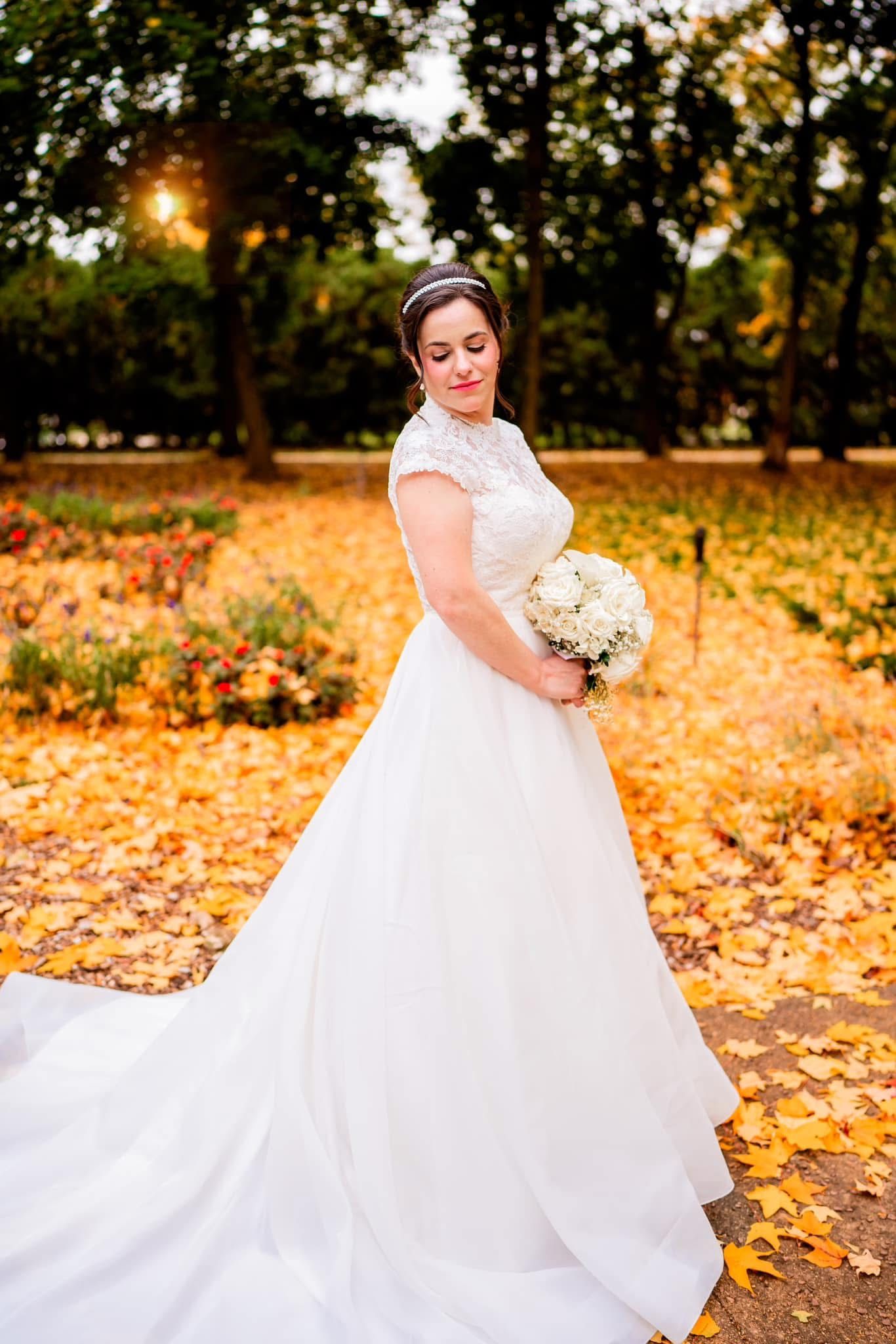 A bride in a white lace gown holds a cream bouquet while standing on fallen autumn leaves in a park at sunset.