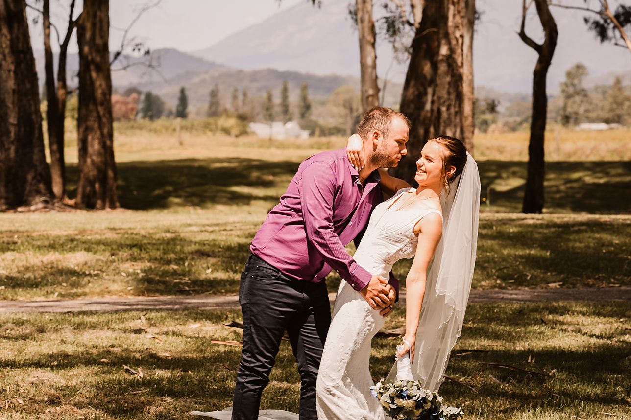 A couple shares an intimate moment in an outdoor setting with mountains and sunlight filtering through trees.