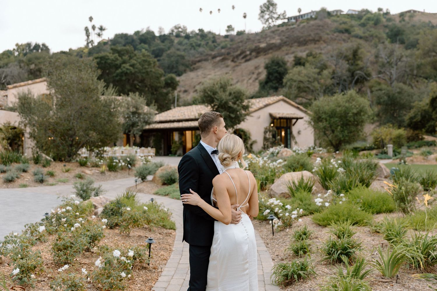 A couple shares a moment together at a rustic outdoor venue surrounded by desert landscaping and hills.