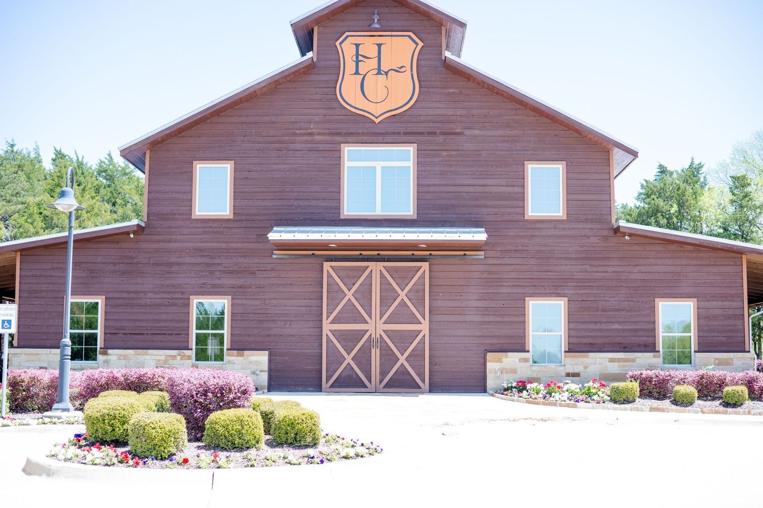 Rustic brown barn building with large sliding doors and decorative shield emblem features landscaped flower beds at entrance.