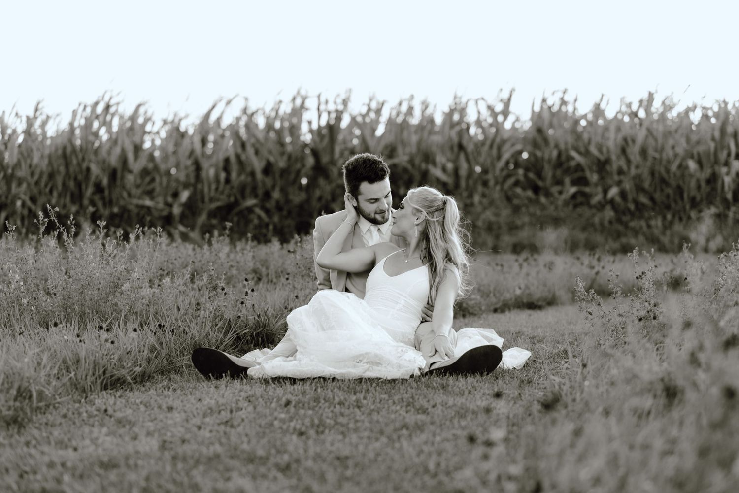 A romantic black and white photo of a couple sitting together in a field near tall grass.