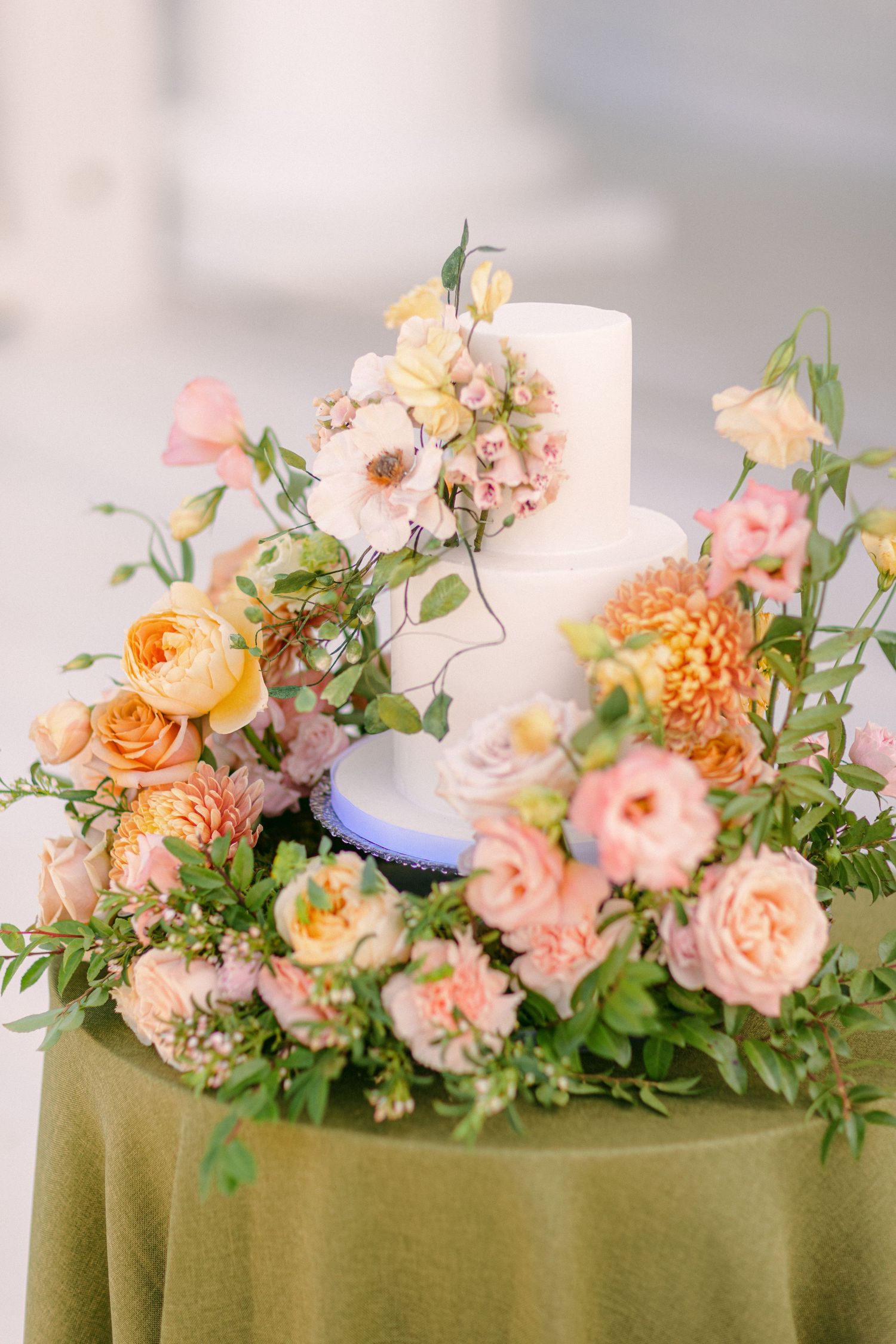A white wedding cake adorned with peach and coral flowers sits on a sage green tablecloth with lush floral arrangements.
