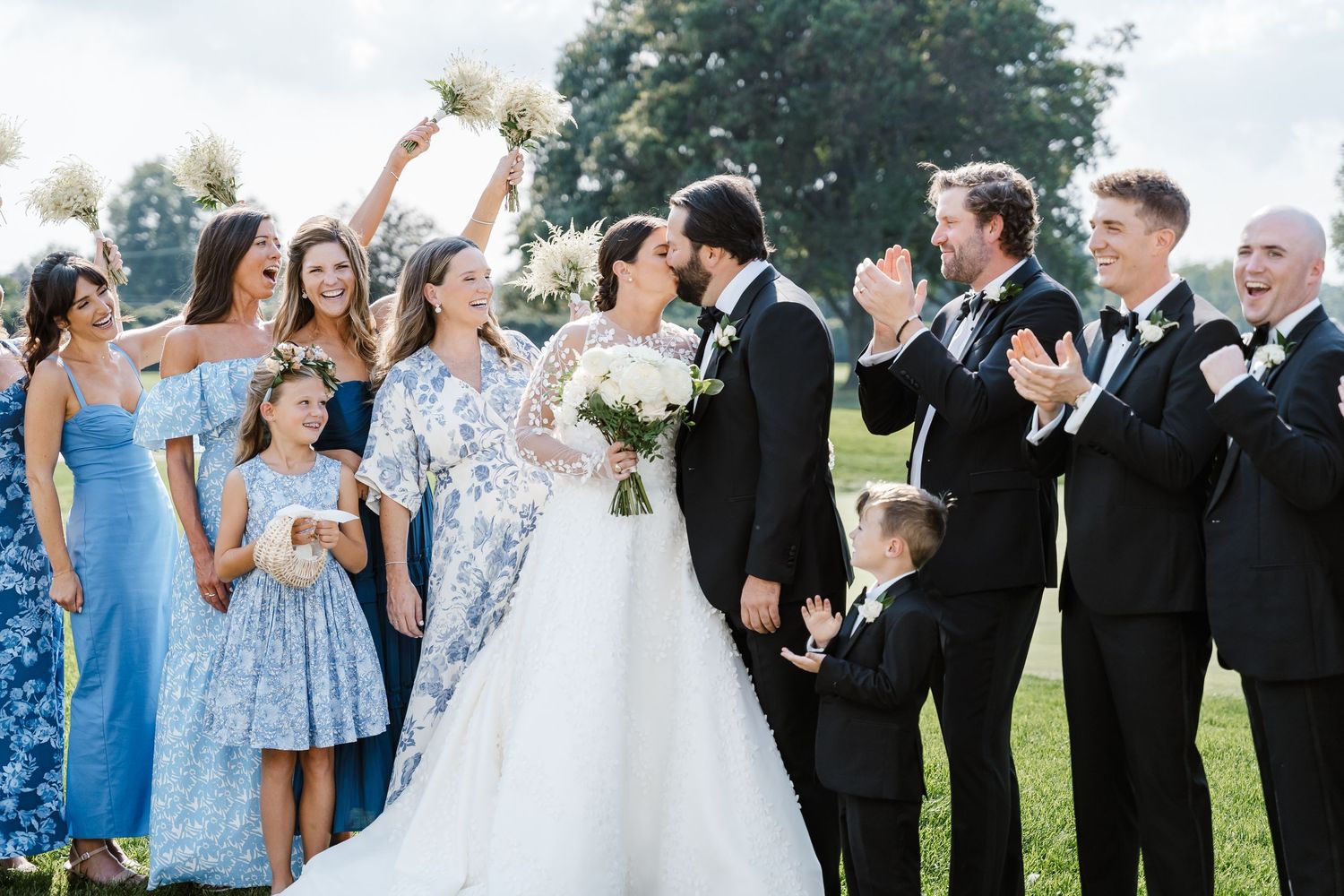Two people in formal attire share a tender moment while holding orange and white floral bouquets in a meadow.