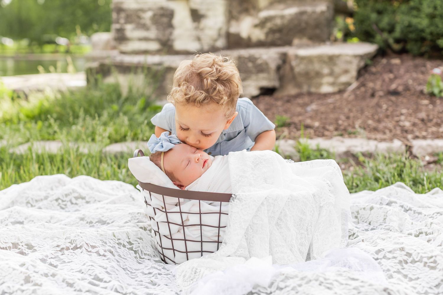 Baby in light blue outfit looks at white blanket while leaning over wire basket outdoors.